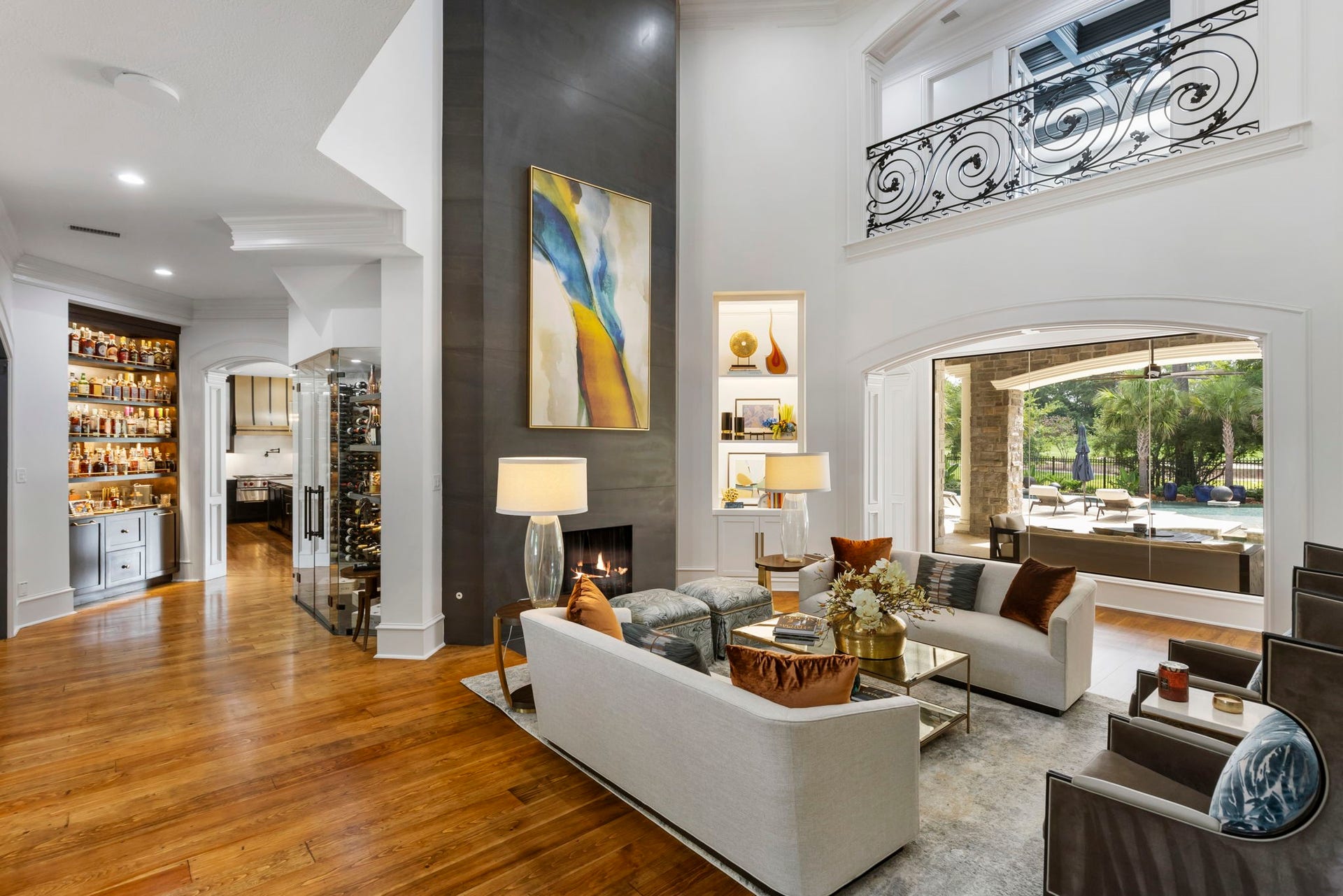 Formal living room centered on a two-story cast stone fireplace wall, with an iron-railed catwalk above & walls of glass opening to the pool & golf course beyond.