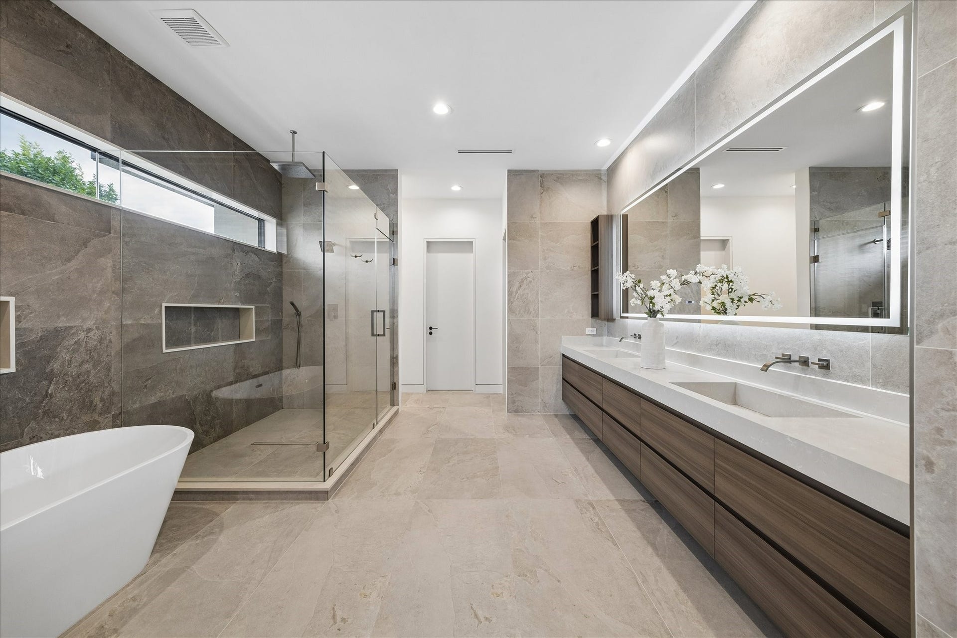 This bathroom features a modern design with a spacious walk-in glass shower, a freestanding bathtub, and a long double vanity with ample storage. The large mirror and soft lighting create an elegant atmosphere, complemented by neutral-toned tiles for a sleek, contemporary look.