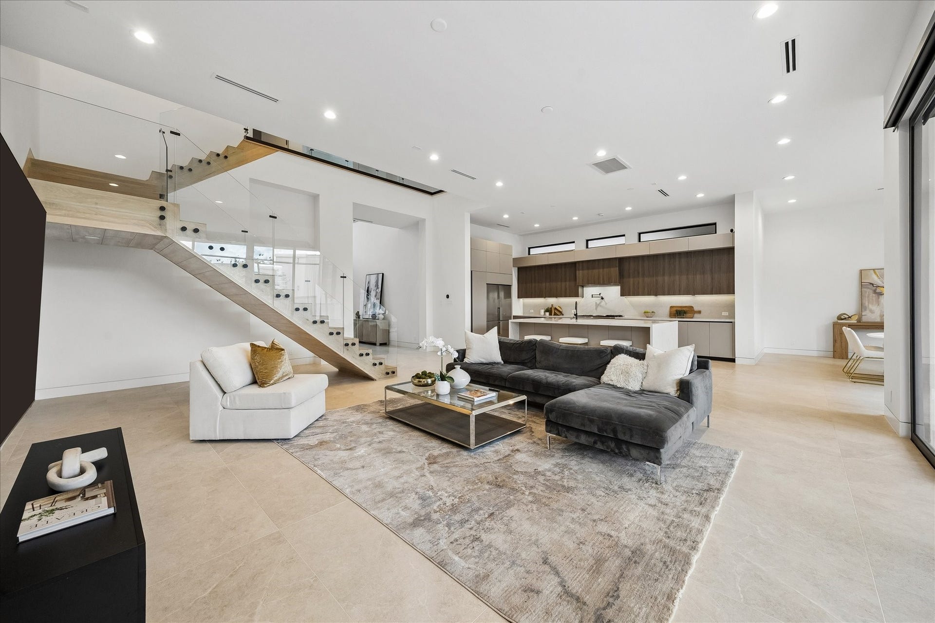 Modern, open-concept living space features a sleek staircase with glass railing, a spacious seating area and a stylish kitchen. Natural light floods in through expansive windows, creating a bright and inviting atmosphere.