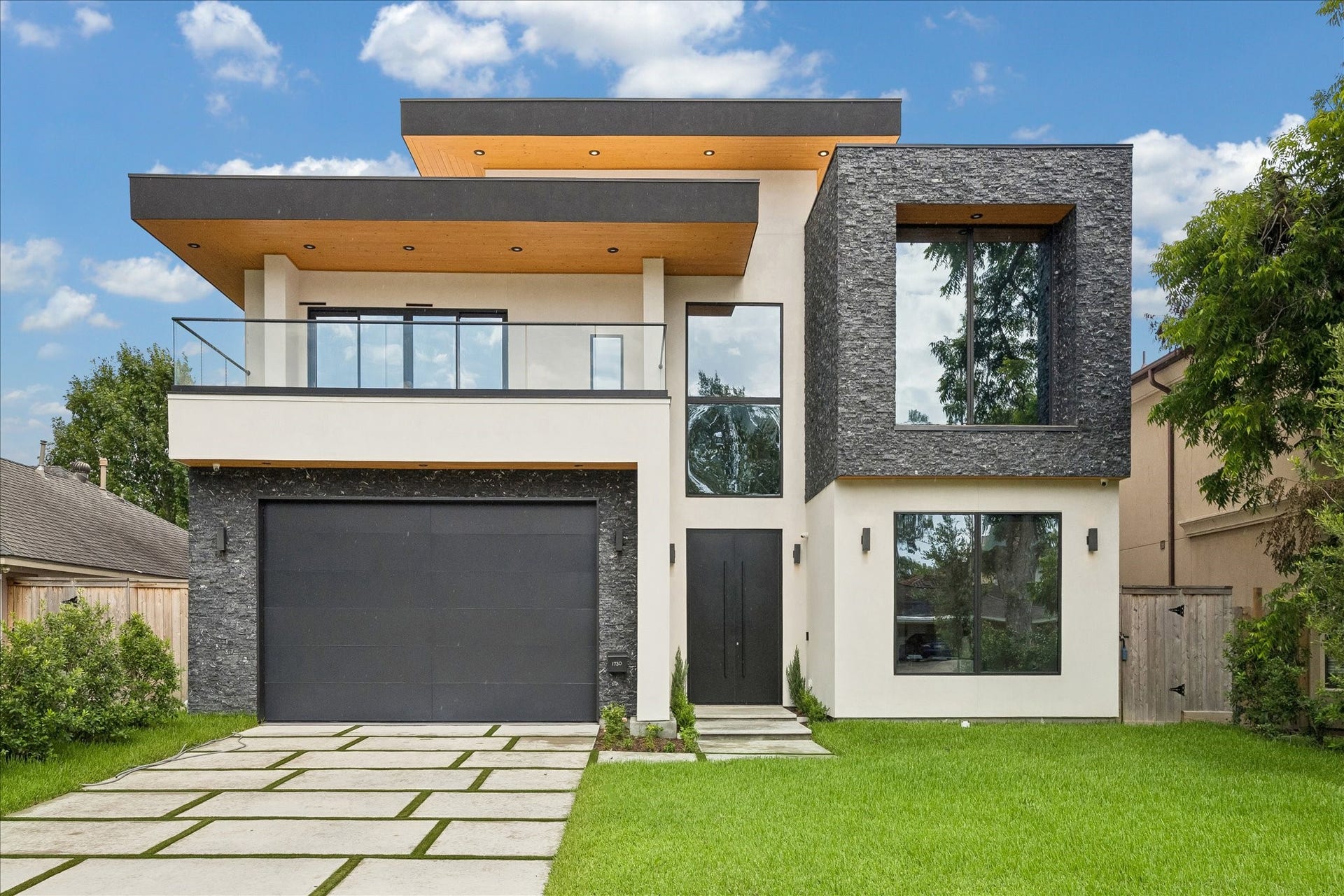 This modern two-story home features a sleek design with a large garage, expansive windows, and a well-maintained lawn. The exterior combines white walls with dark stone accents, offering a contemporary and stylish aesthetic.