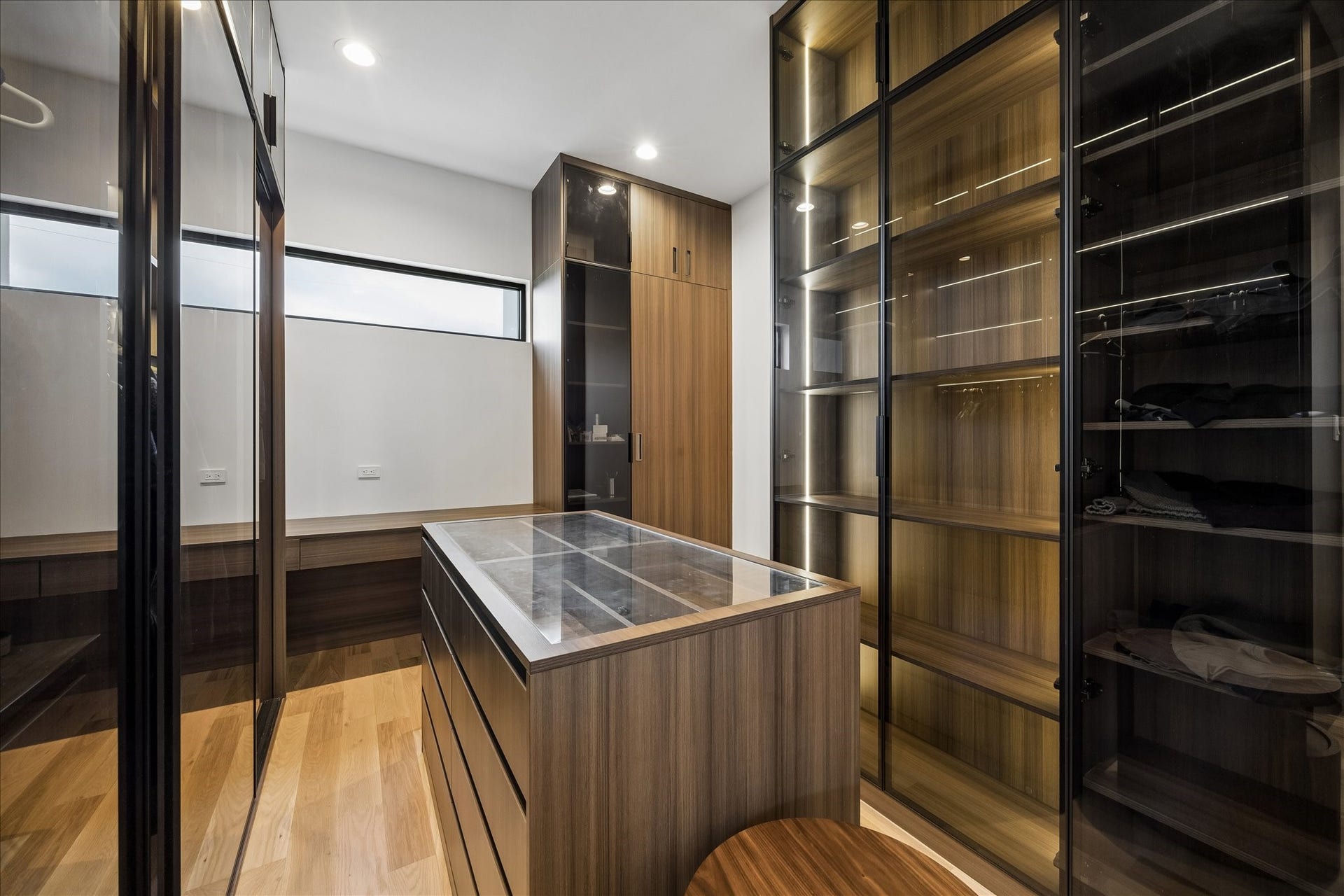 Two walk-in closets have sleek glass-front cabinets, LED lighting, ample shelving, and a central island with storage drawers.