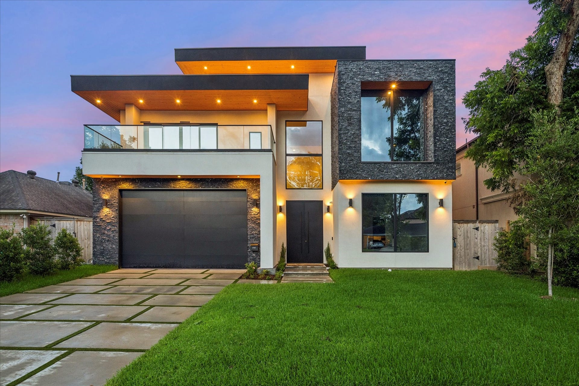 This modern home features a striking facade with clean lines, large windows, and a spacious balcony. The well-lit exterior is complemented by sleek stone accents and a manicured lawn, providing an inviting curb appeal.