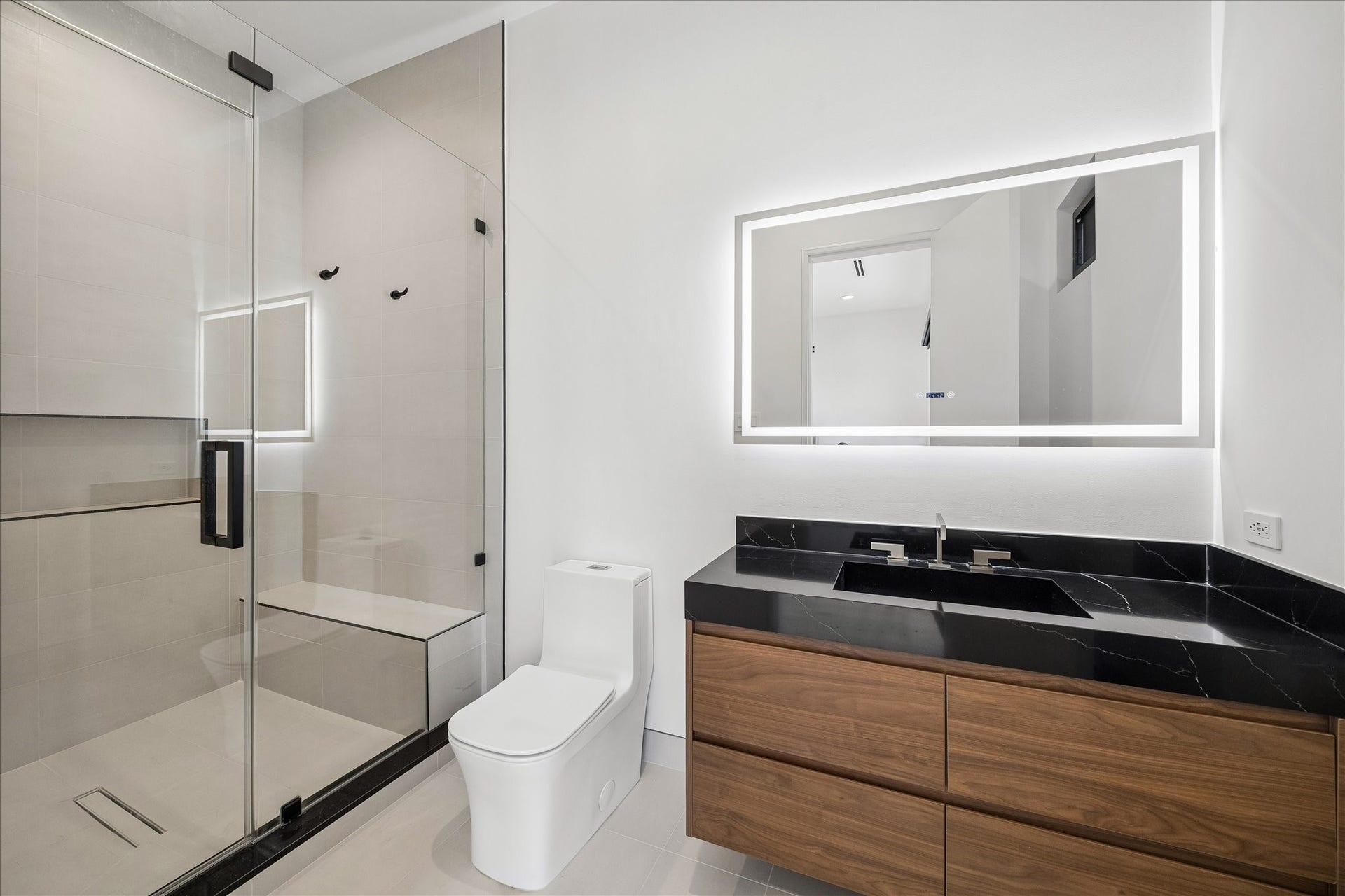 Modern bathroom features a sleek design with a glass-enclosed shower, a stylish vanity with a black countertop. The large, LED illuminated mirror adds a touch of elegance and enhances the space.