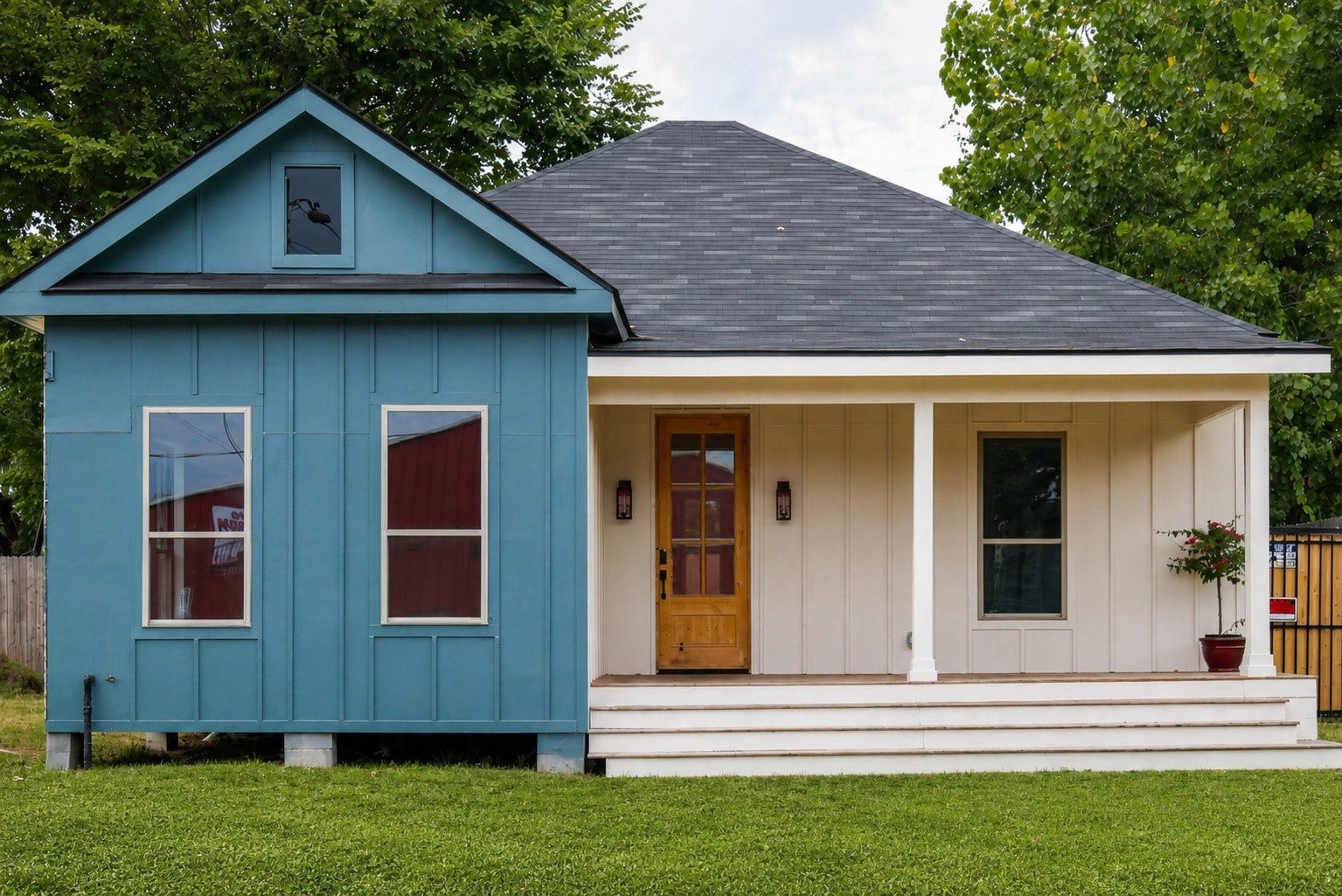 This charming home features a unique two-tone exterior with a bright blue and neutral color scheme. It has a welcoming front porch, a modern design, and is surrounded by a spacious yard with mature trees and a wooden fence, offering both privacy and curb appeal.