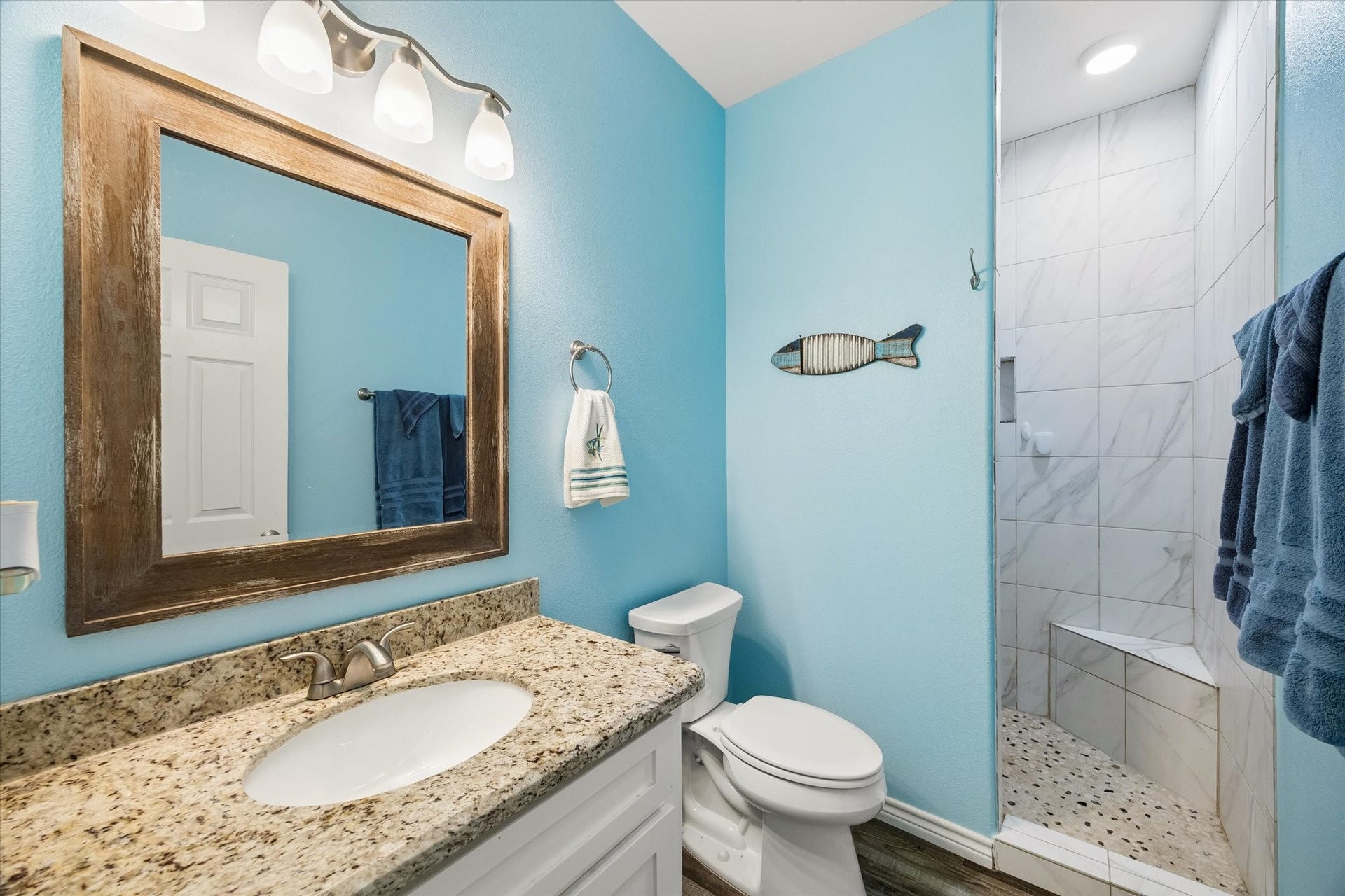 161 Four Master Road, Freeport, TX 77541 Charming bathroom with a soothing blue color scheme, featuring a granite countertop, rustic wood-framed mirror, and a stylish tiled shower with a built-in bench.