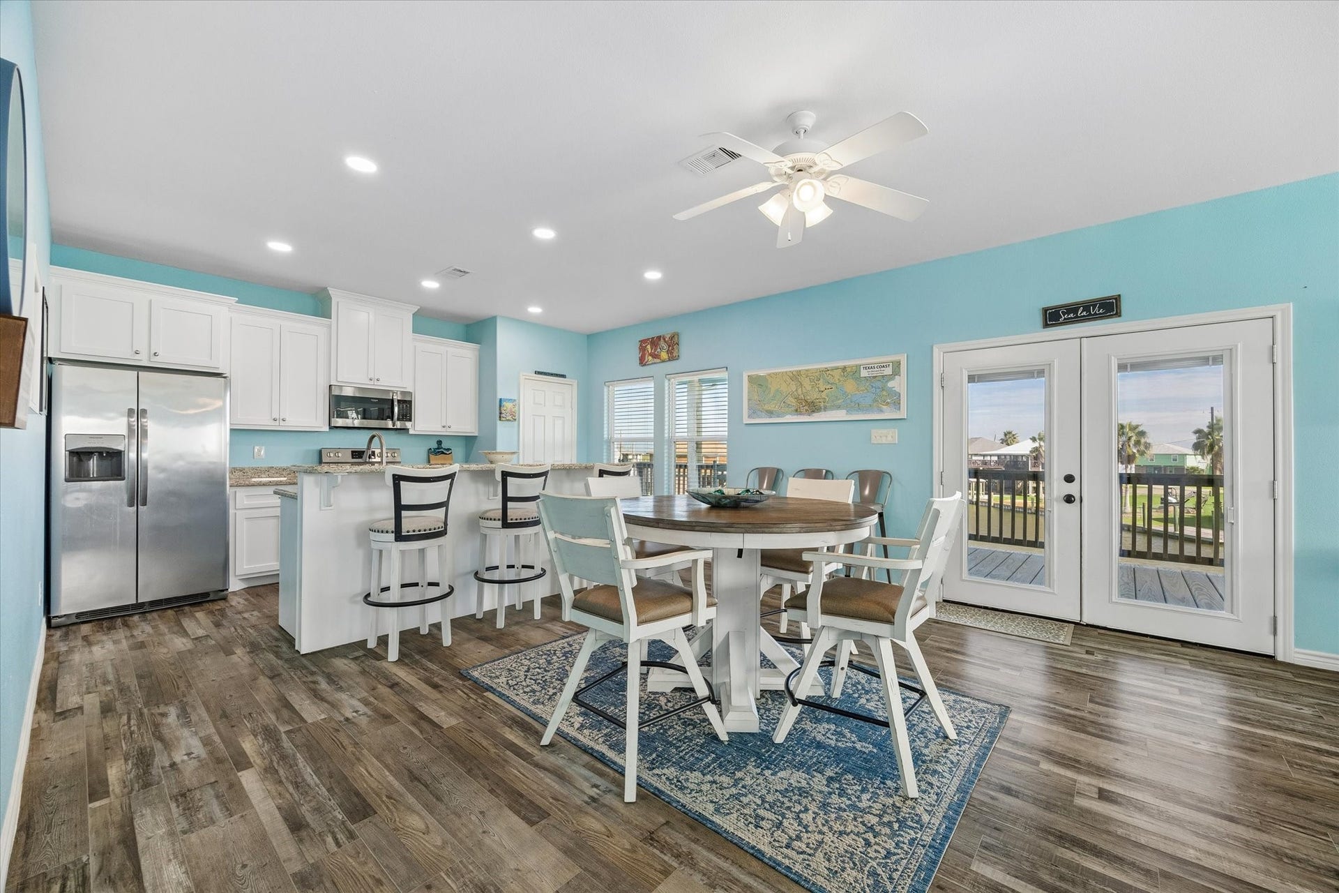 161 Four Master Road, Freeport, TX 77541 Experience the perfect blend of style and functionality in this bright and airy kitchen-dining area, featuring white cabinetry, a sleek stainless steel fridge, and a central island, all enhanced by modern lighting, cheerful blue walls, and French doors leading to scenic outdoor views.
