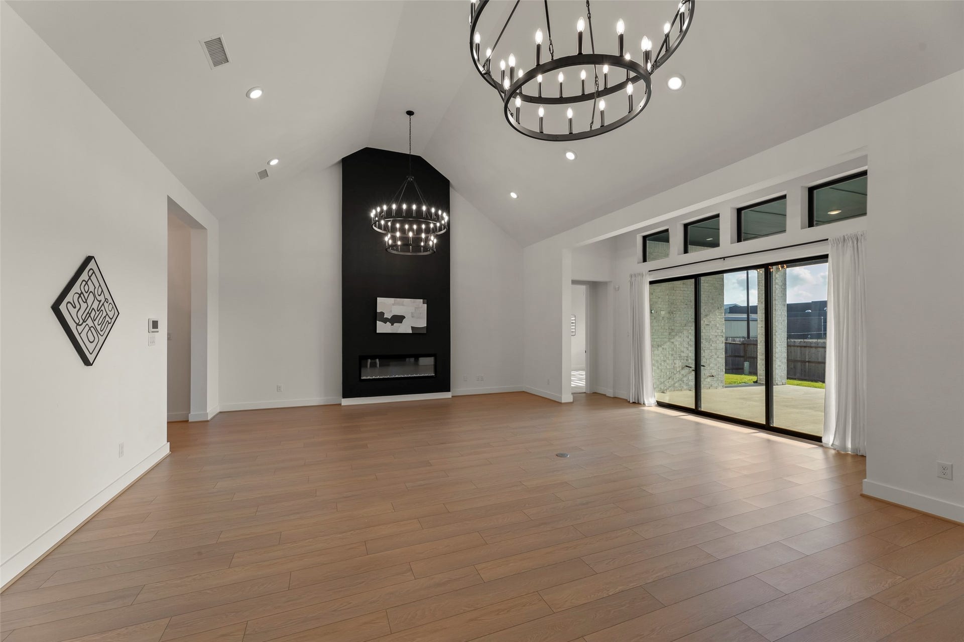 2015 Jade Manor Drive, Rosenberg, TX 77469 Vaulted ceilings, elegant chandeliers, and a modern black accent wall featuring a fireplace. Expansive windows and sliding glass doors offer ample natural light and access to a patio.