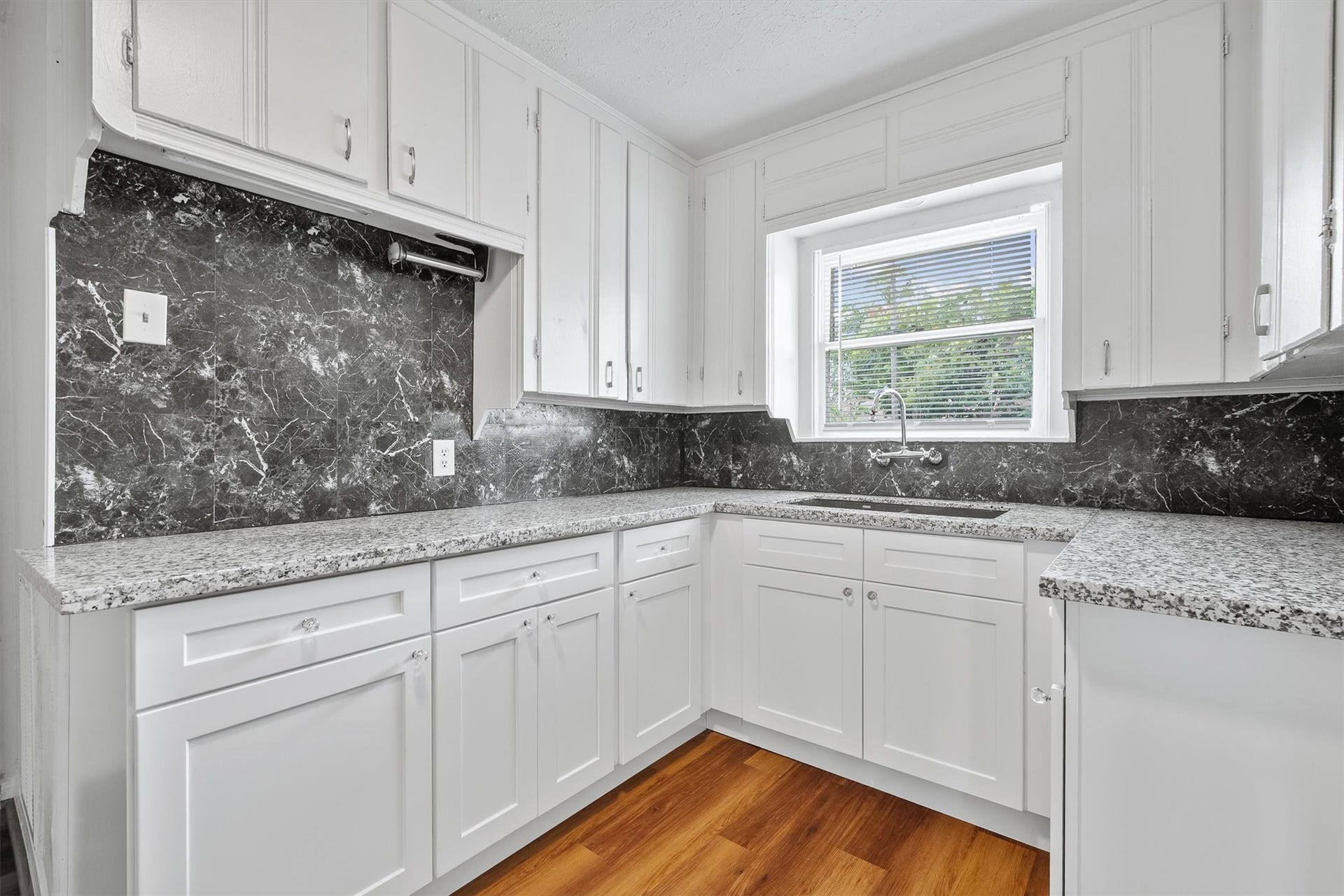 6630 Maybank Drive, Houston, TX 77055 Bright kitchen featuring sleek white cabinetry, elegant granite countertops, and a striking marble backsplash, complemented by warm hardwood floors and ample natural light from the large window.