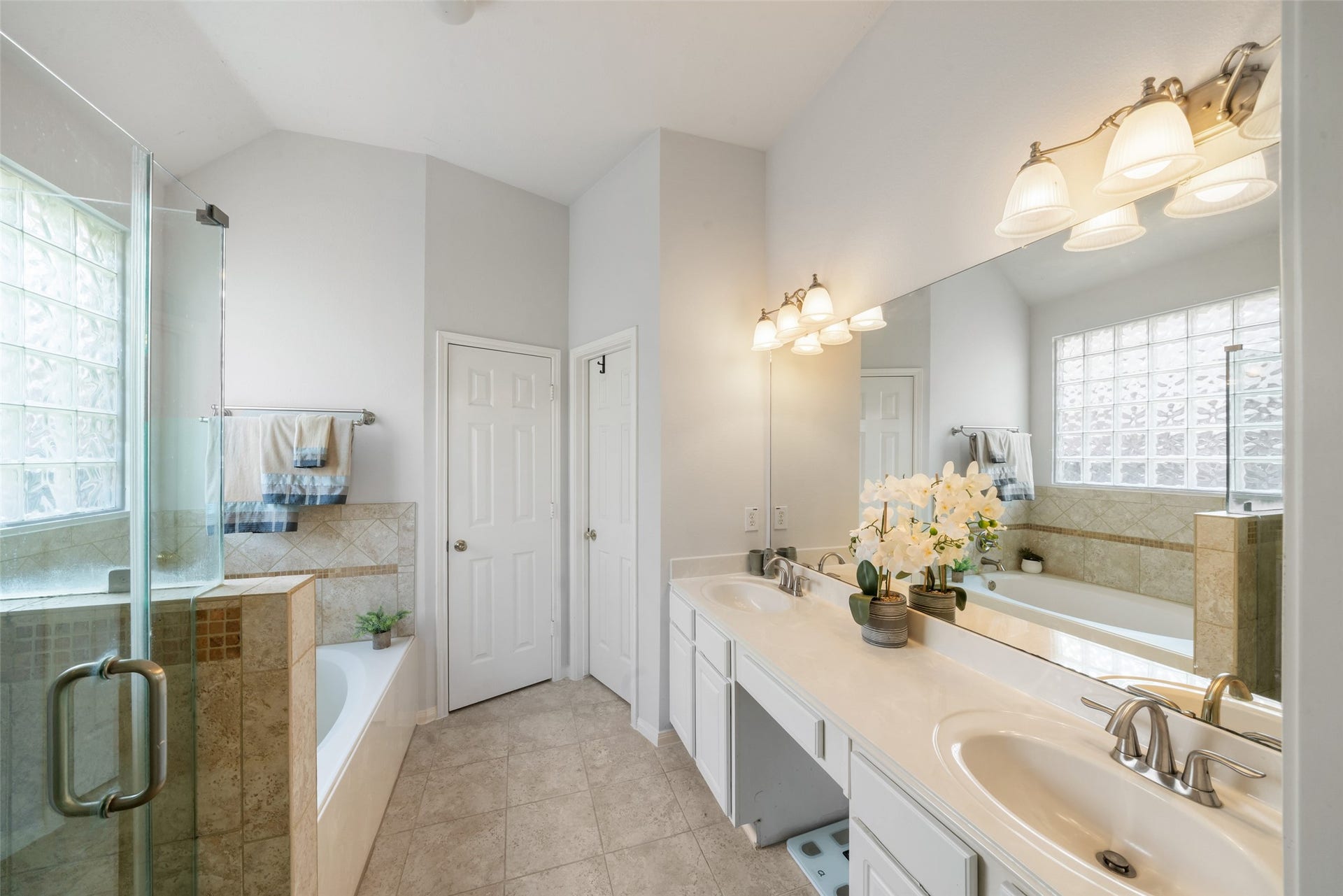 3607 Parkshire Drive, Pearland, TX 77584 Elegant bathroom featuring dual sinks, a glass-enclosed shower, and a spacious soaking tub, enhanced by abundant natural light from glass-blocked windows. Doors lead to a water closet on the left and a large walk-in closet on the right.