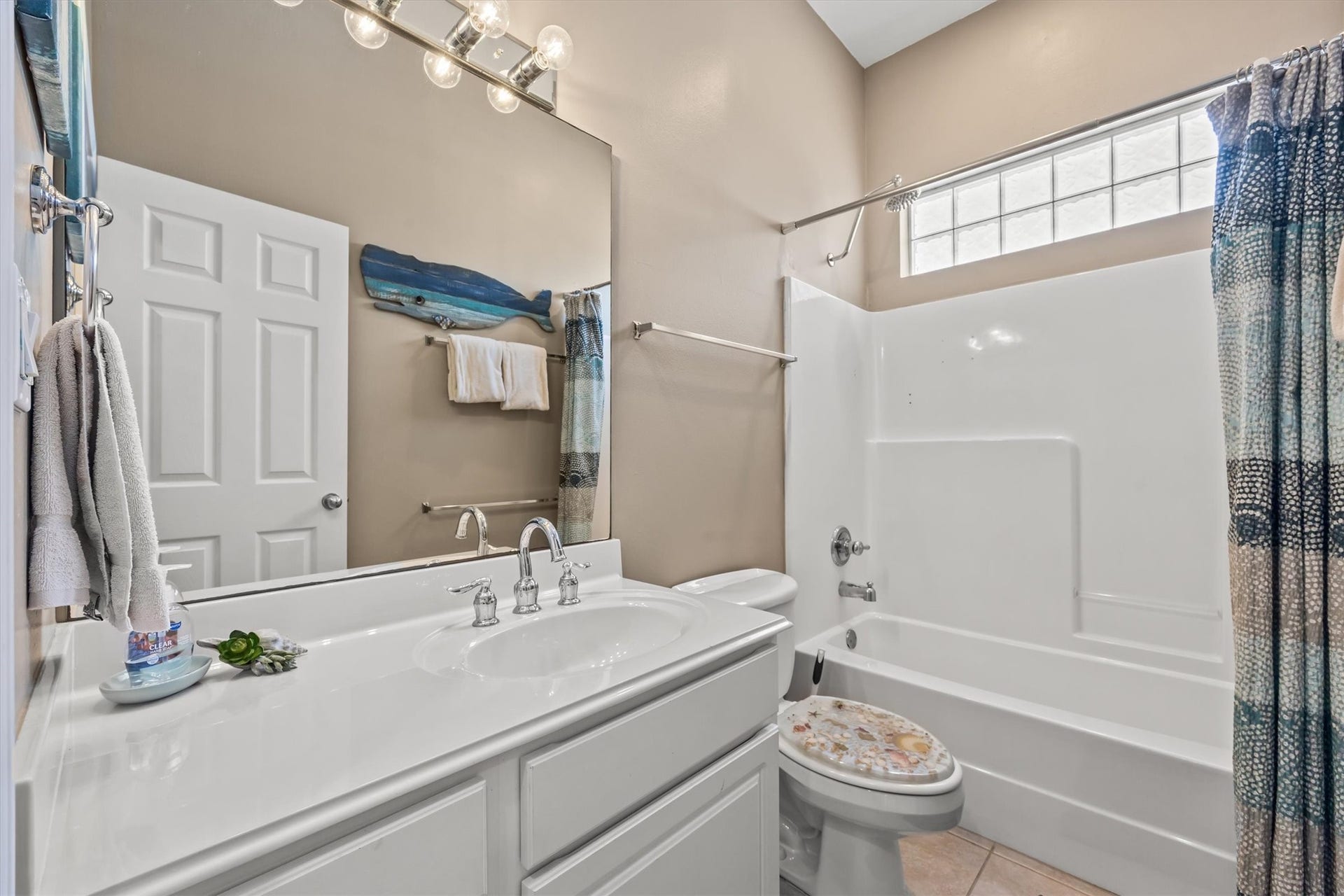 21519 San Luis Pass Road, Galveston, TX 77554 This bathroom features a clean, modern design with a large mirror, ample lighting, and a white vanity. The space includes a bathtub with a shower, accented by a colorful curtain. Natural light enters through a glass block window, enhancing the room's bright and airy feel.