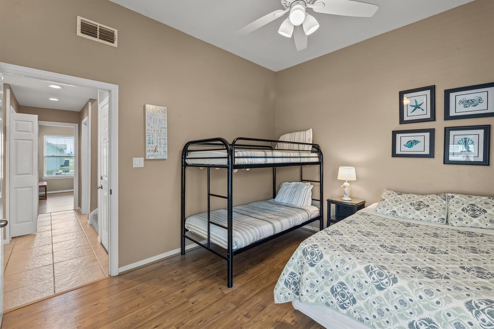 21519 San Luis Pass Road, Galveston, TX 77554 This cozy bedroom features a bunk bed and a queen-sized bed, with a ceiling fan for comfort. The space is warmly painted and decorated with coastal framed art. A hallway leads to an additional bedroom.