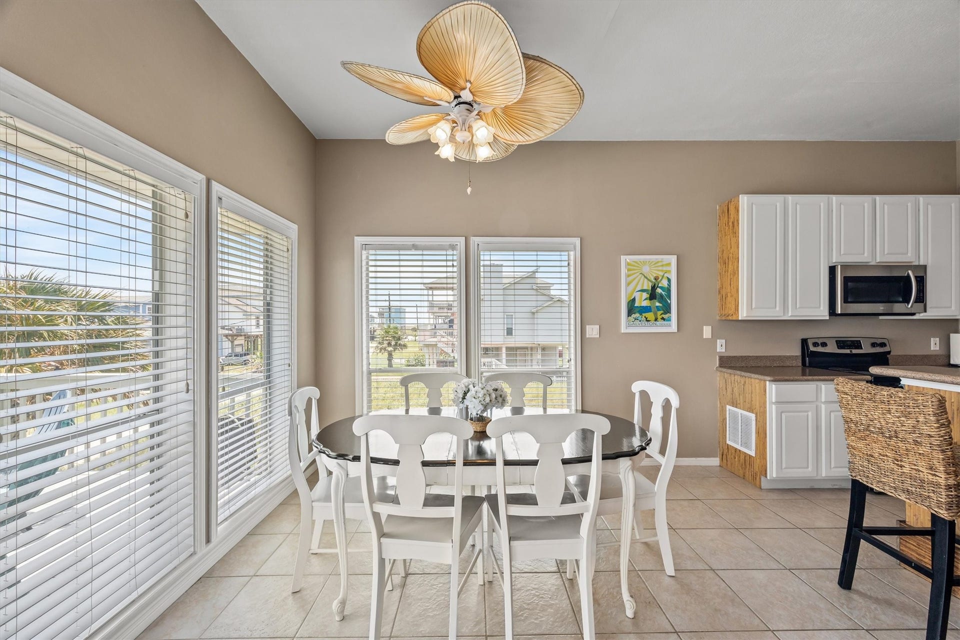 21519 San Luis Pass Road, Galveston, TX 77554 Bright dining area with large windows offering ample natural light. Features a large table with white chairs for family groups, a unique ceiling fan, and a view of a sunny day or a beautiful sunset. Conveniently connected to the modern kitchen with white cabinetry and stainless steel appliances.