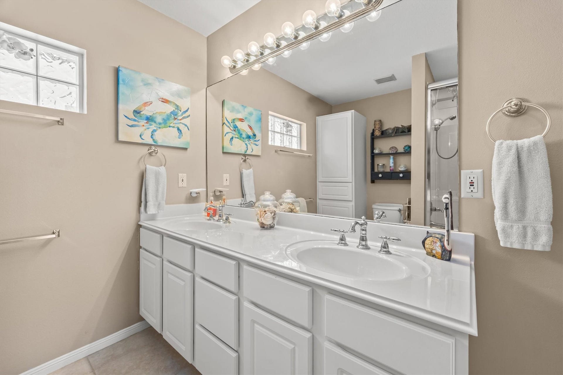 21519 San Luis Pass Road, Galveston, TX 77554 This bathroom features a double vanity with sleek white cabinetry, a large mirror with great lighting, and neutral-toned walls. It includes decorative artwork, a glass block window for natural light, and a shower area. The space is modern and inviting.