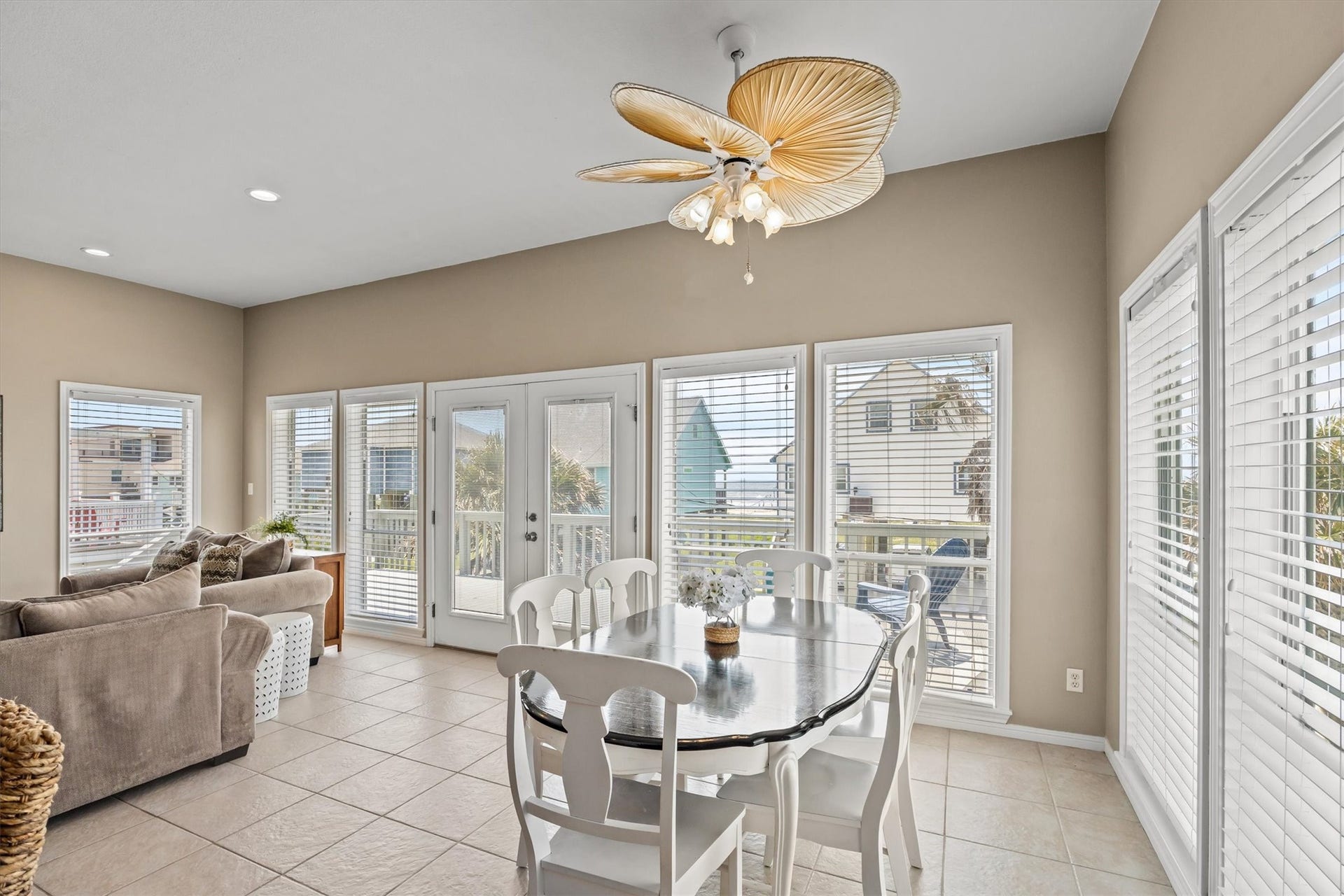 21519 San Luis Pass Road, Galveston, TX 77554 This bright and airy living space features large windows with white blinds, a stylish ceiling fan, and an open layout that includes a cozy sitting area and a dining table. The space opens to the deck with views of sunny blue sky and the beach offering a welcoming and comfortable atmosphere.