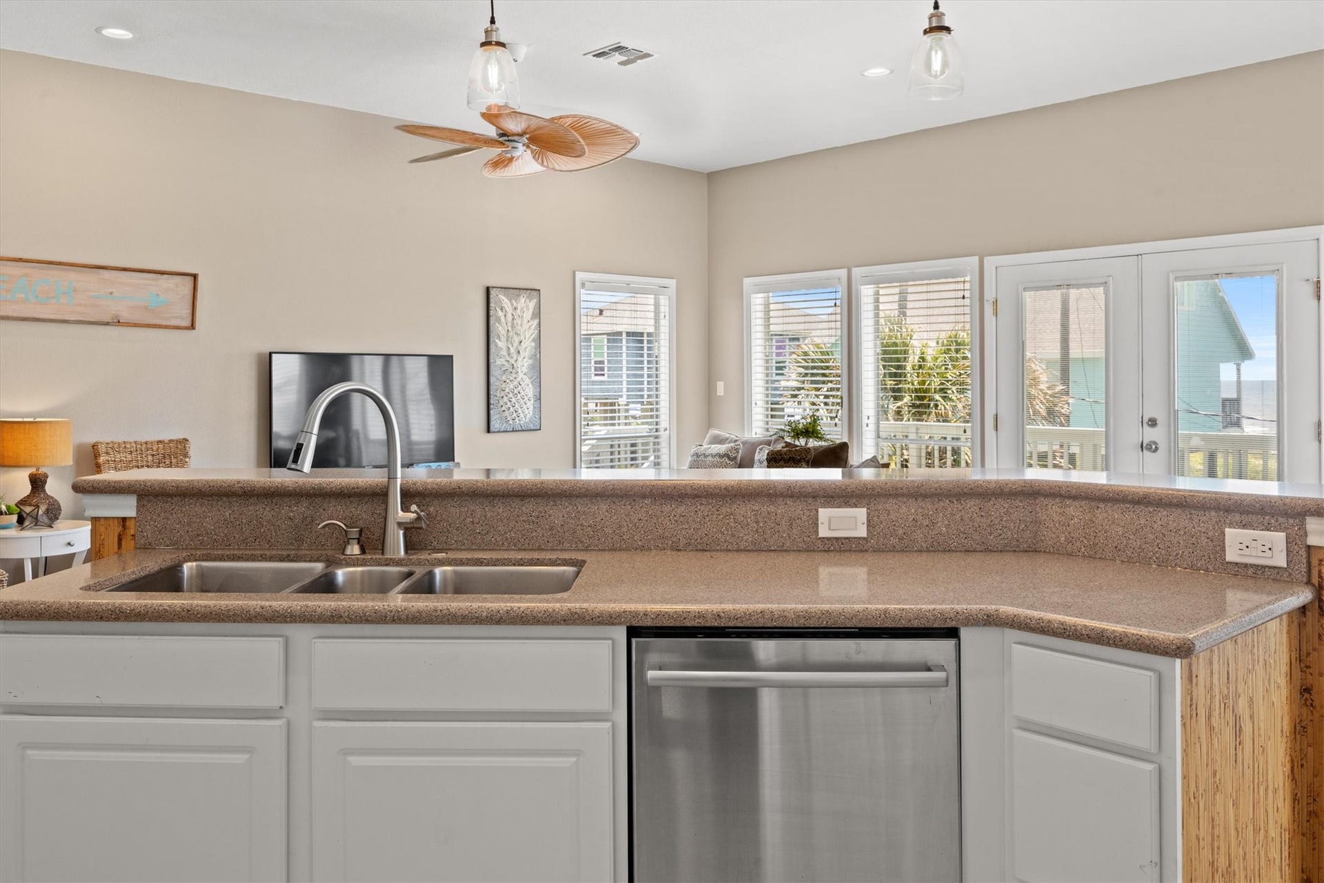 21519 San Luis Pass Road, Galveston, TX 77554 This photo shows bright, open kitchen overlooking the living area. There's a modern kitchen island with a double sink and stainless steel dishwasher backed by the roomy bar for casual eating. The space is well-lit with natural light from large windows and glass doors that lead to an outdoor area. The room features neutral tones and a ceiling fan, creating a welcoming atmosphere.