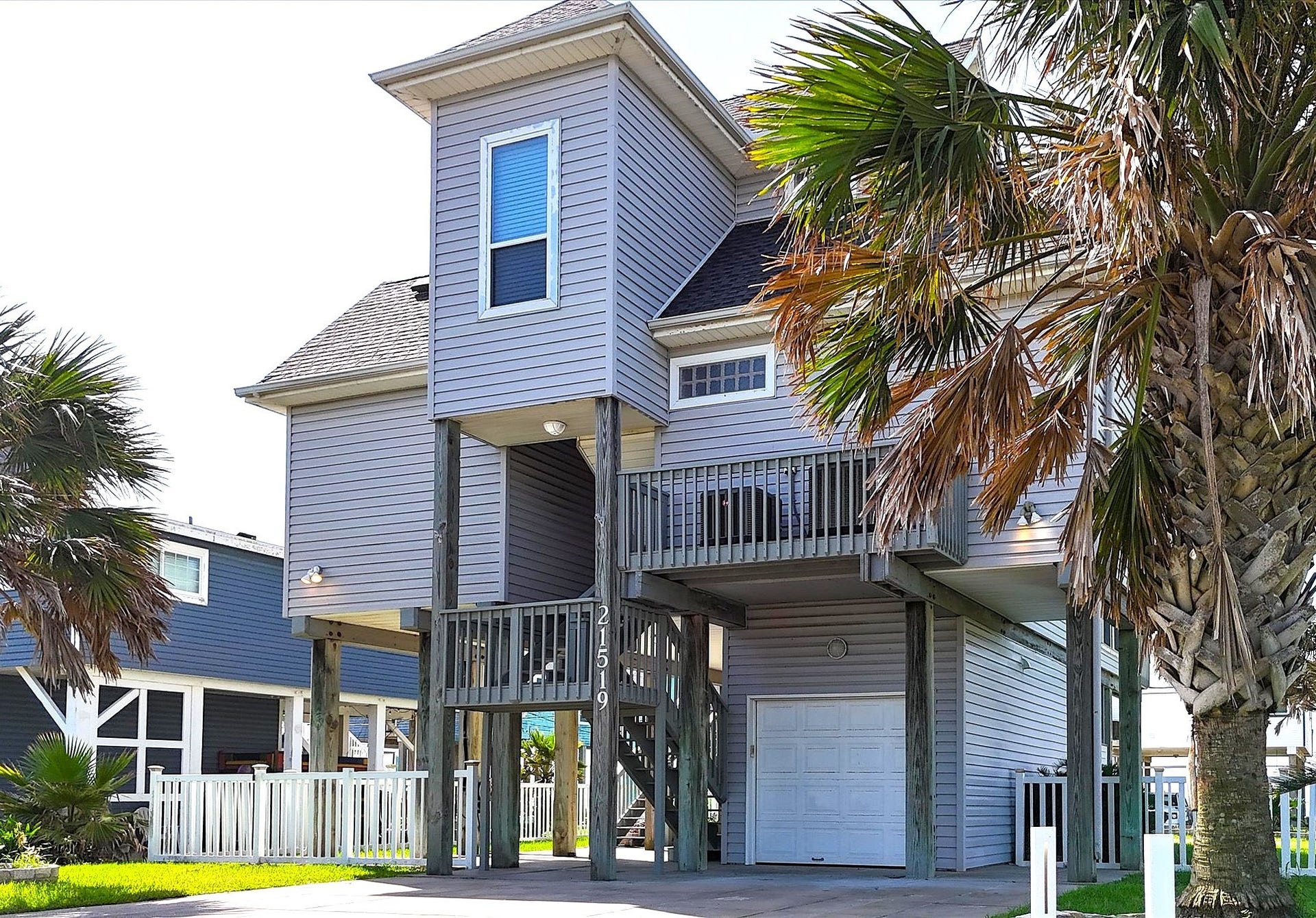 21519 San Luis Pass Road, Galveston, TX 77554 This photo shows the underside of the raised coastal home with a wooden staircase leading to the main floor, surrounded by a grassy yard and palm trees. The space beneath the house is open with concrete flooring, providing ample parking or storage.