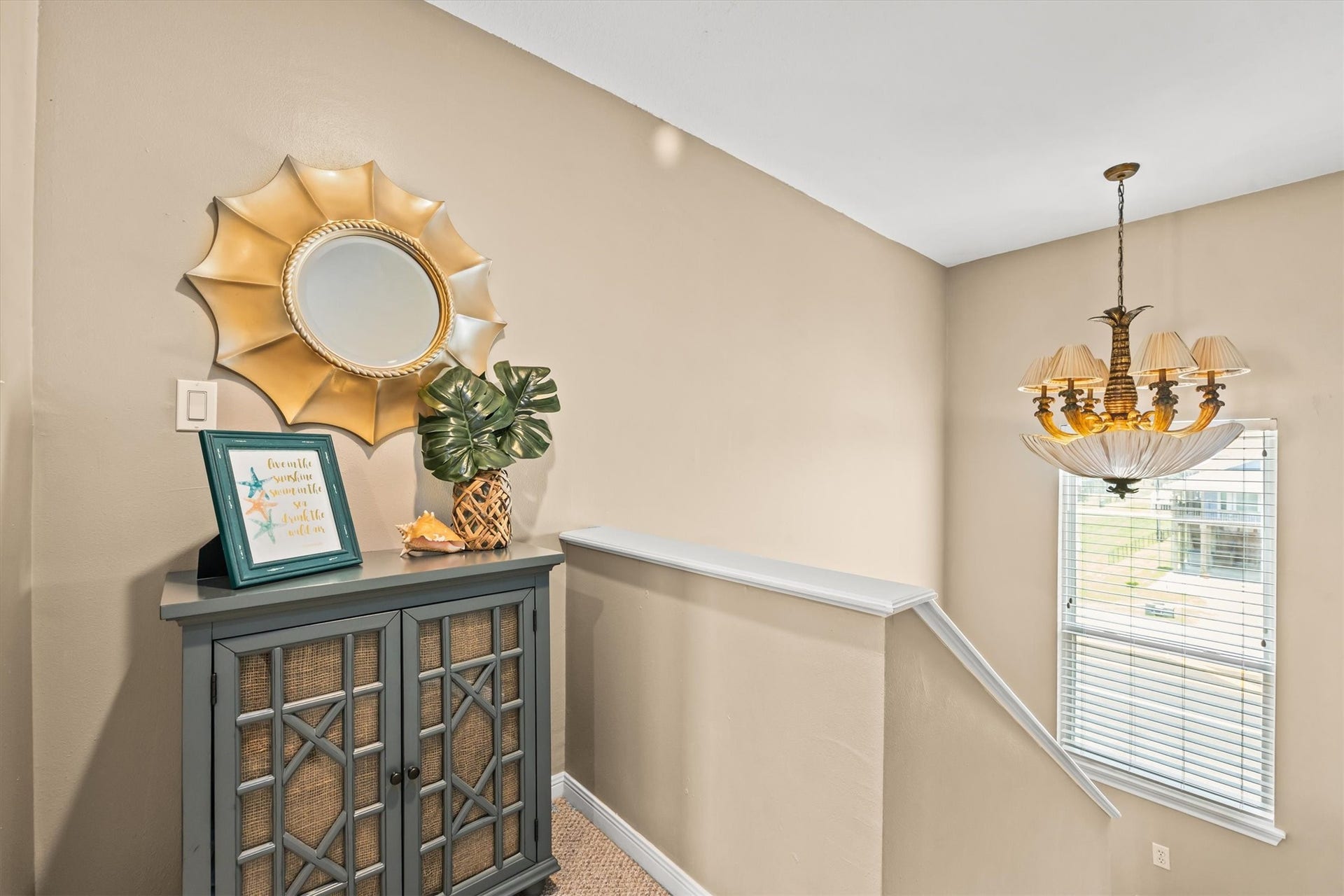 21519 San Luis Pass Road, Galveston, TX 77554 This photo shows an open hallway landing outside of the primary suite featuring a cabinet topped. A stylish sunburst mirror hangs above, and a classic chandelier illuminates the space. A window with blinds provides great natural light.