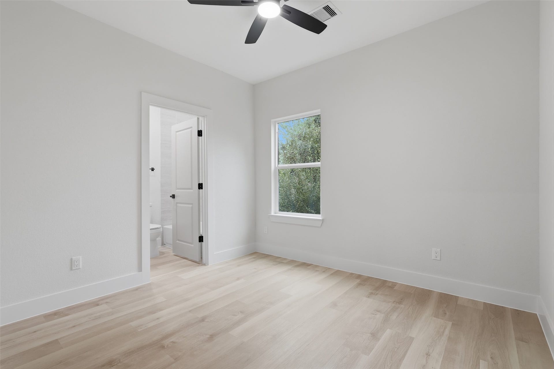 3201 & 3205 Berry Street, #A/B, Houston, TX 77004 Bright and airy bedroom featuring light wood like LVP flooring, a ceiling fan, and a window with scenic views. Convenient ensuite bathroom access enhances functionality.