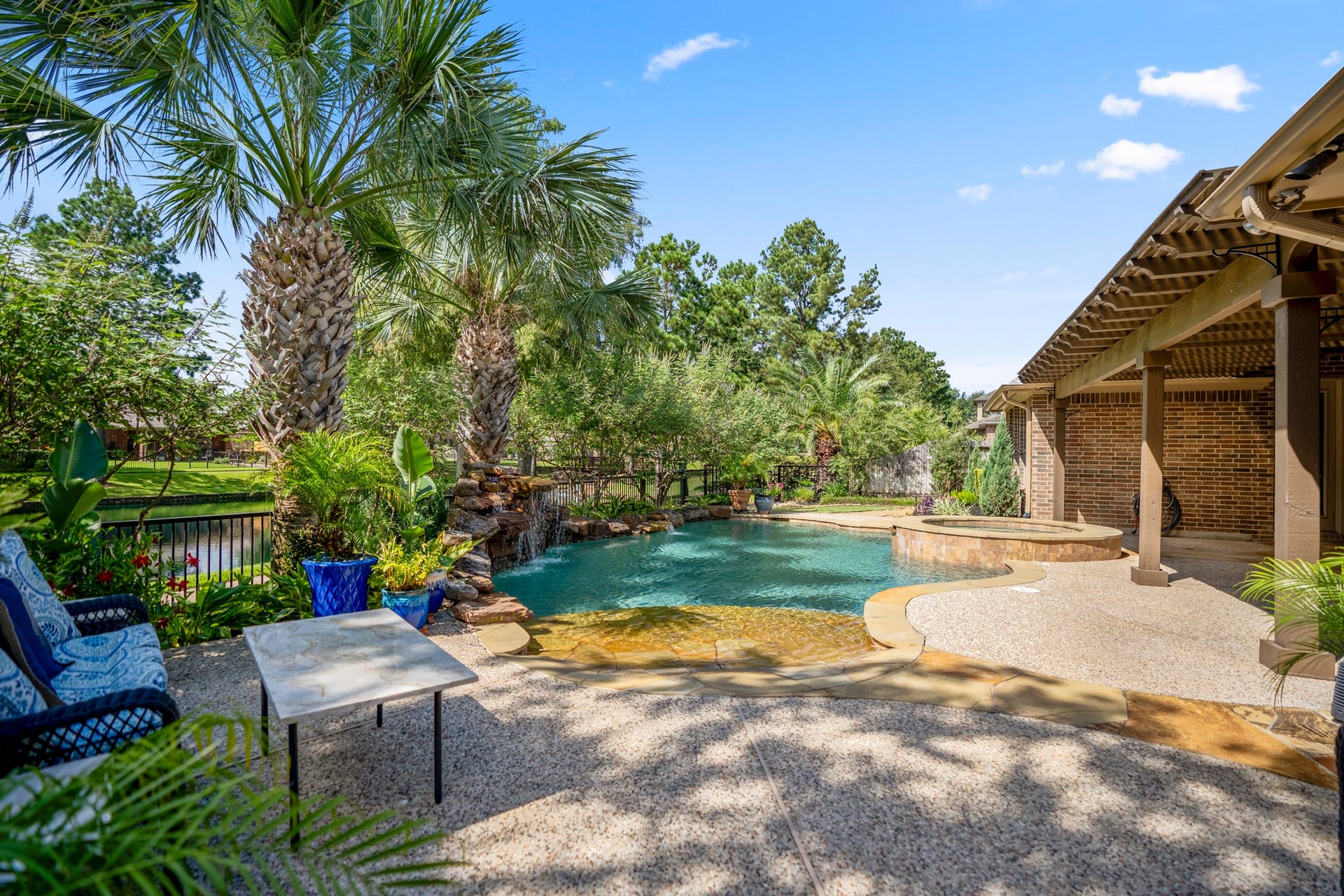2407 Hendricks Lakes Drive, Spring, TX 77388 Poolside from the far end of the backyard, this view captures the beach-style pool entry, raised spa, and water beyond the yard for a fuller sense of the outdoor setting. The covered pergola stretches along the right side of the home, while palm trees and layered landscaping wrap the space with a relaxed, resort-style feel made for slow evenings, weekend swims, and time outside.