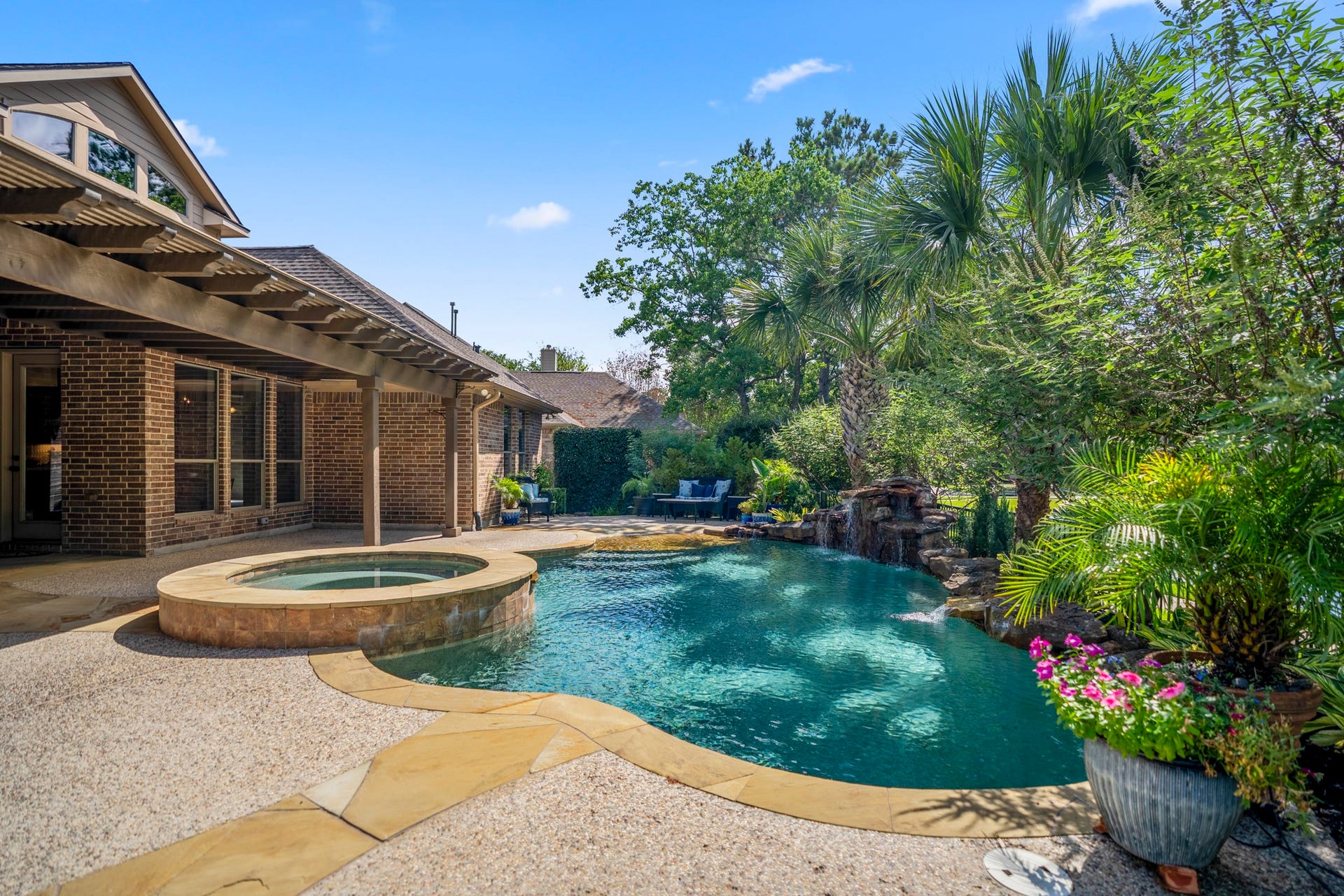 2407 Hendricks Lakes Drive, Spring, TX 77388 Poolside, the backyard feels wrapped in greenery, with the raised spa, curved pool, and surrounding landscaping creating a private-feeling place to relax outdoors. The covered pergola stretches along the back of the home on the left, while palm trees and layered shrubs soften the view and set up the kind of evening backdrop the sellers loved most.