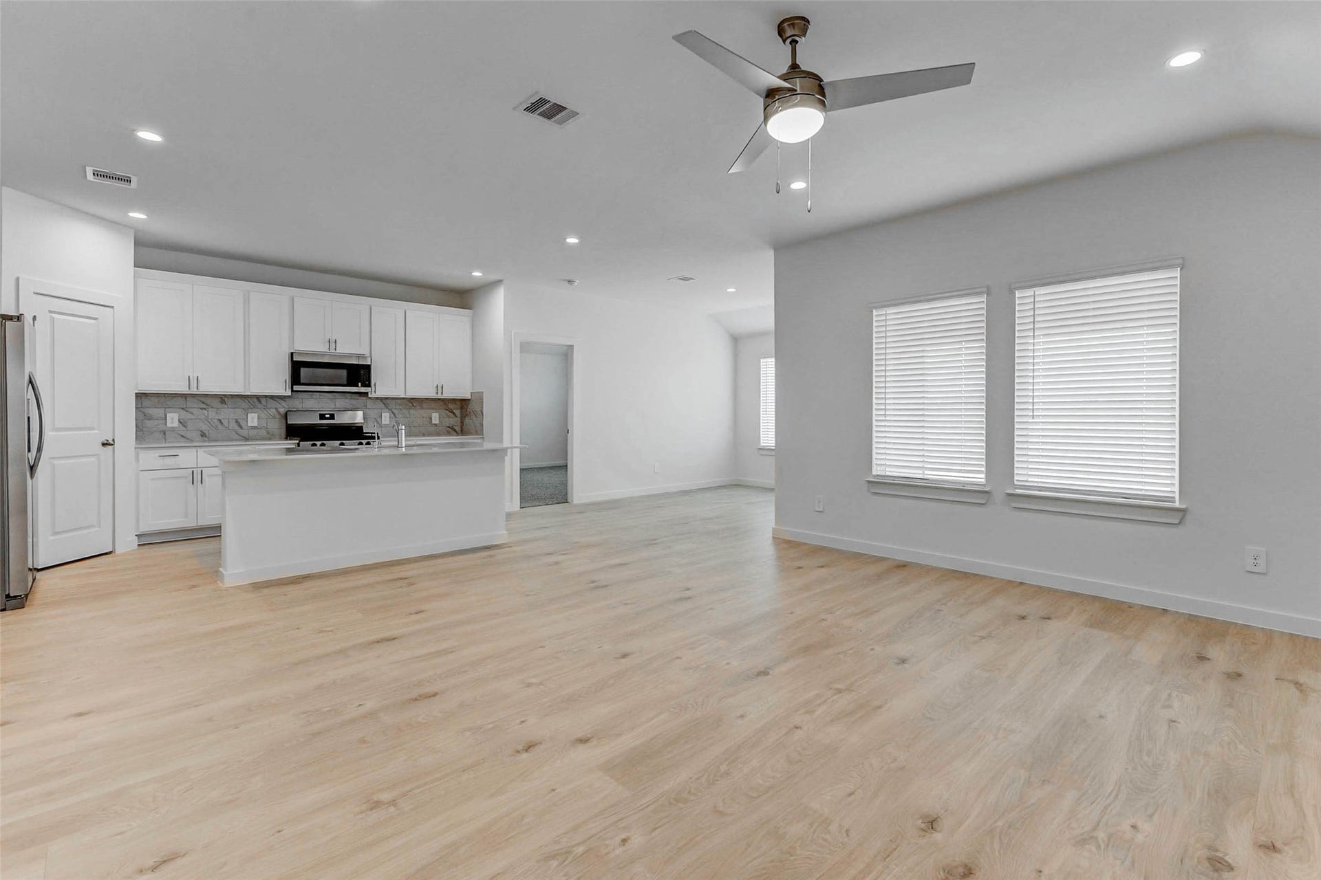 30307 Lighthouse Bluff Lane, Fulshear, TX 77441 Modern, open-concept kitchen and living area. It features light wood flooring, white cabinetry, a central island with a sink, stainless steel appliances, and ample natural light from large windows. A ceiling fan is centrally located, complementing the clean and bright aesthetic.