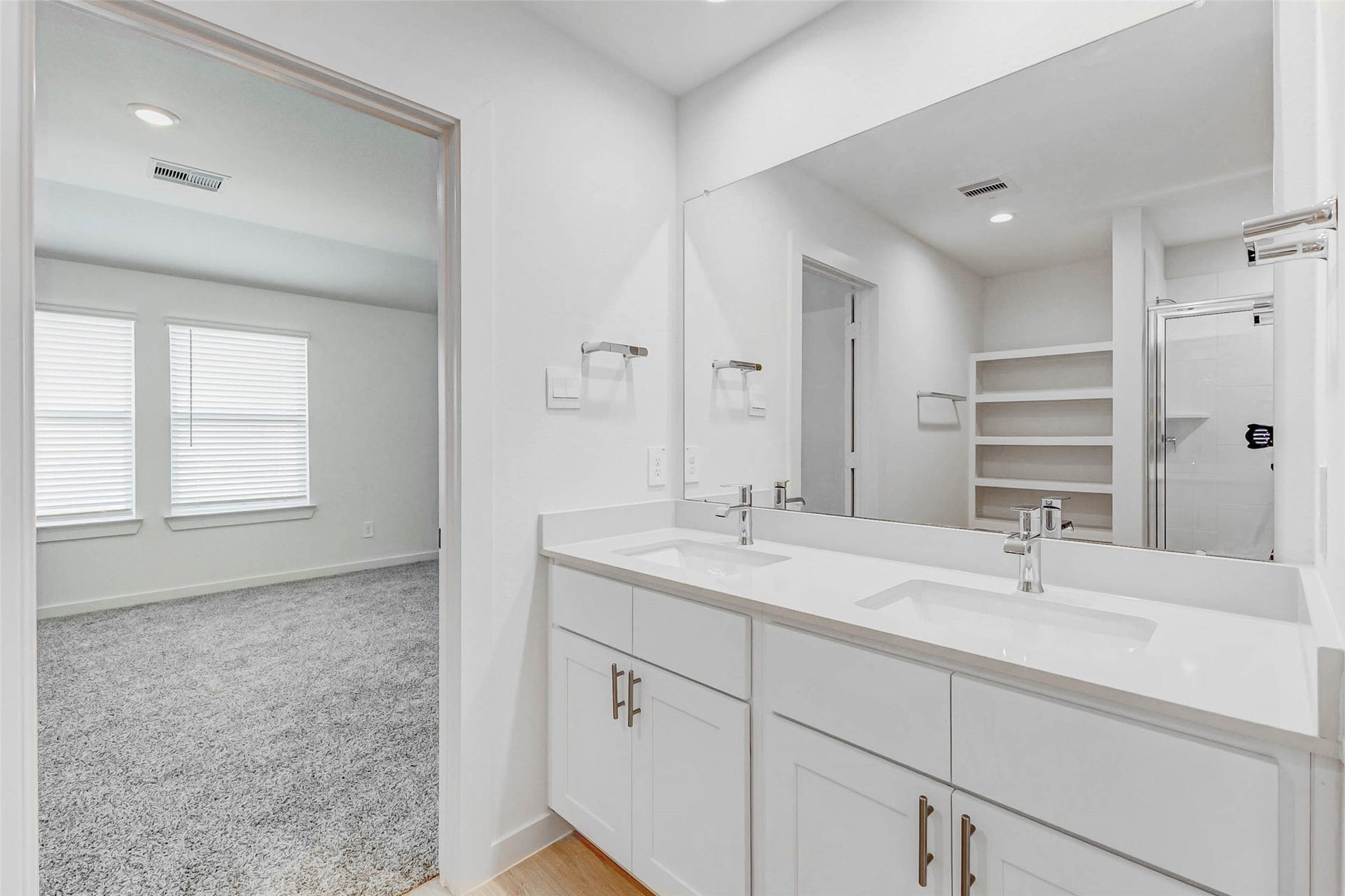 30307 Lighthouse Bluff Lane, Fulshear, TX 77441 Modern bathroom with dual sinks, white cabinetry, and a spacious mirror. Adjacent carpeted bedroom with large windows, and a shower area with built-in shelving. Bright and open layout.