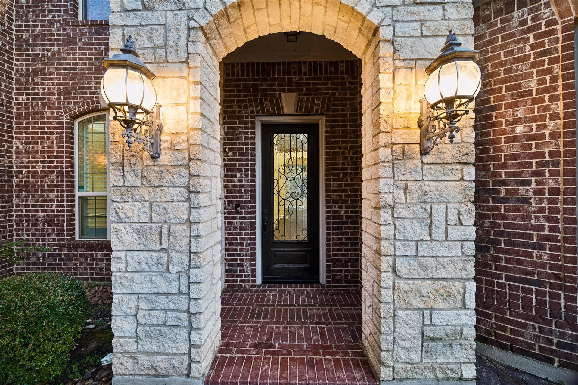 The elegant entry welcomes guests with timeless architectural details, including a custom iron-and-glass front door, warm lantern-style lighting, and a beautiful combination of rich brick and natural stone finishes.