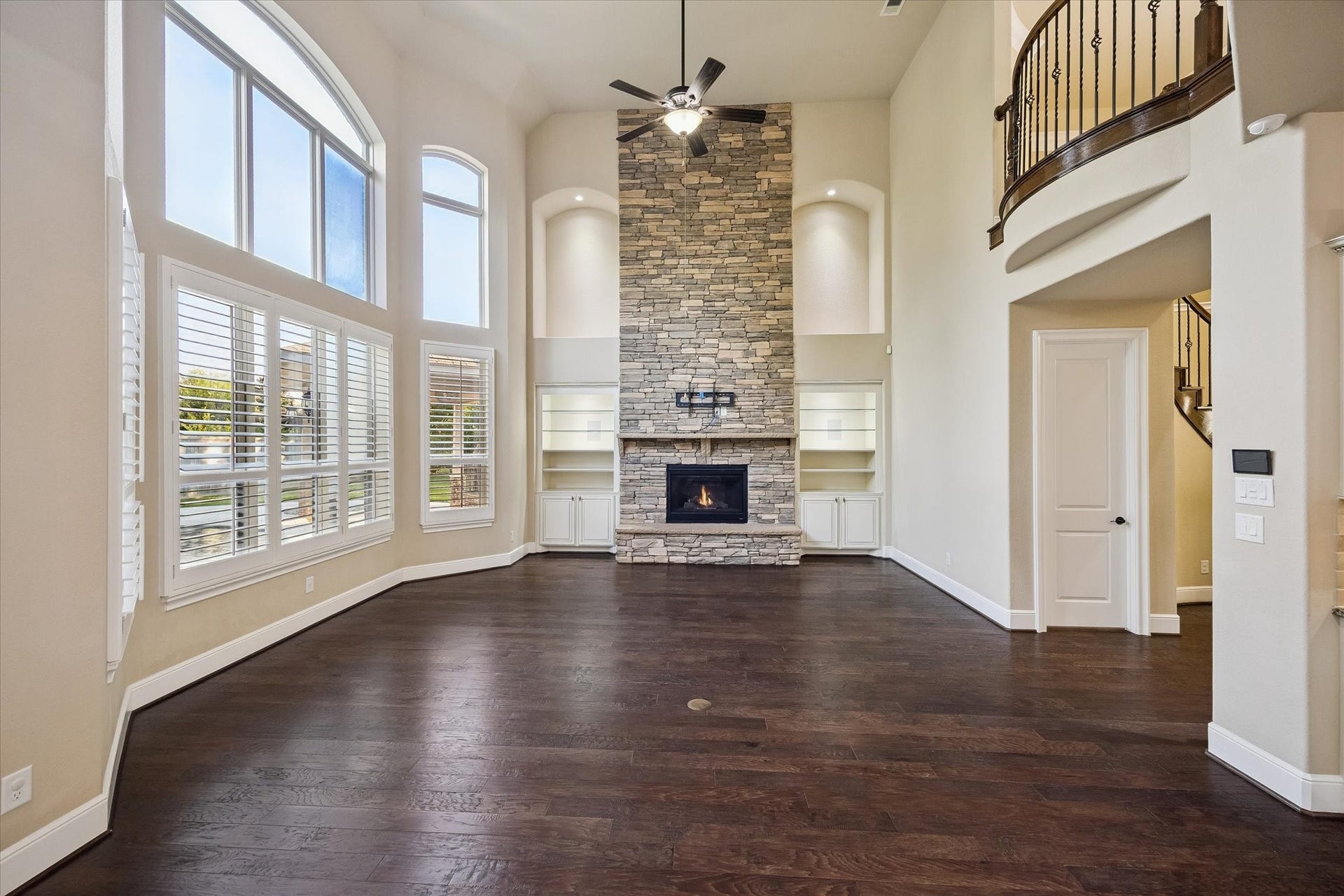 Bathed in natural light, the grand living room features soaring ceilings, rich hardwood floors, expansive windows with plantation shutters, and a striking floor-to-ceiling stone fireplace, creating a warm yet sophisticated gathering space.