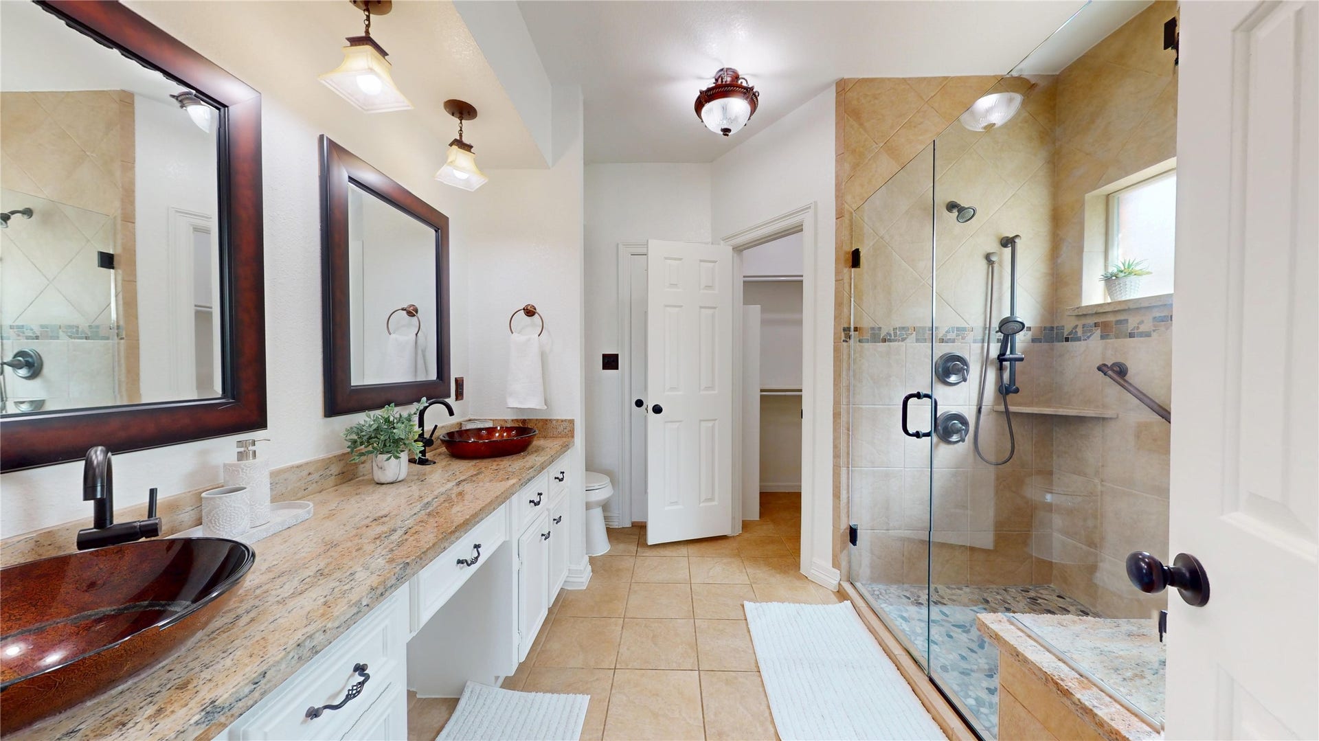 1407 Sherfield Ridge Drive, Katy, TX 77450 1407 Sherfield Ridge Dr, Spacious primary bathroom featuring a long dual-sink vanity with granite countertops, decorative vessel sinks, and large framed mirrors. The oversized walk-in shower offers frameless glass doors, tiled walls, built-in shelving, multiple shower fixtures, and a convenient bench. Neutral tile flooring and warm finishes complete the space.