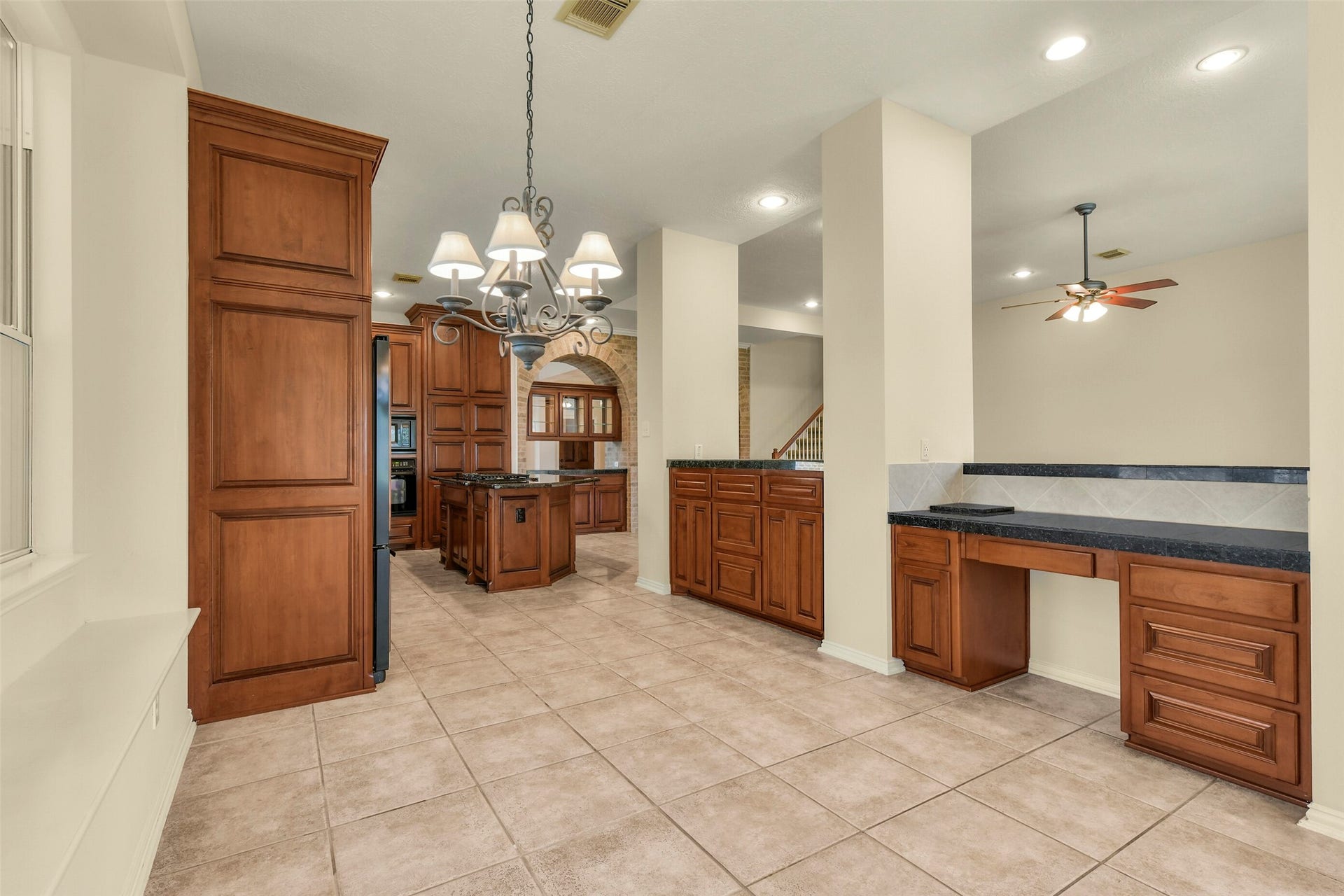 9902 Autumn Lake Trail, Pearland, TX 77584 Spacious kitchen area featuring rich wooden cabinetry, elegant tile flooring, and a convenient built-in desk, seamlessly connected to an open living space with a ceiling fan.