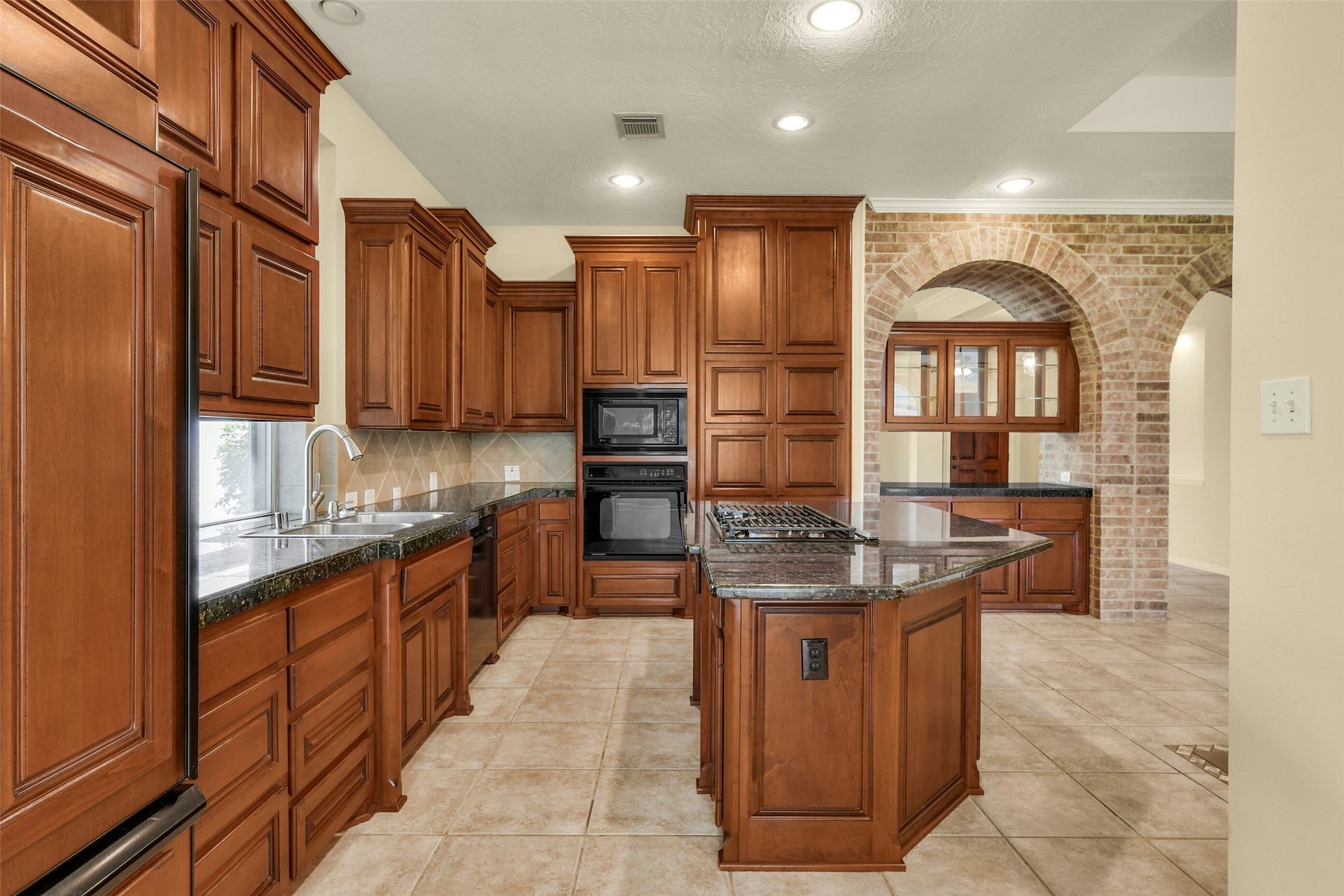 9902 Autumn Lake Trail, Pearland, TX 77584 Elegant kitchen featuring rich wooden cabinetry, a spacious island with a built-in cooktop, and a charming brick archway leading to additional storage space.