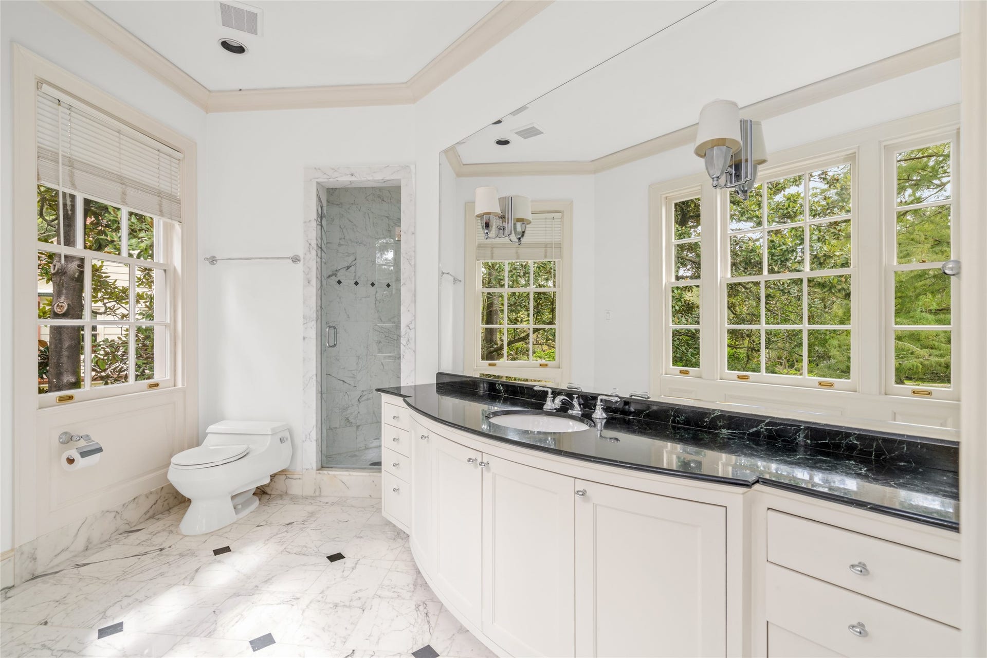 Bright bathroom with a curved vanity, black marble countertops, marble shower and floors, and windows overlooking the front yard.
