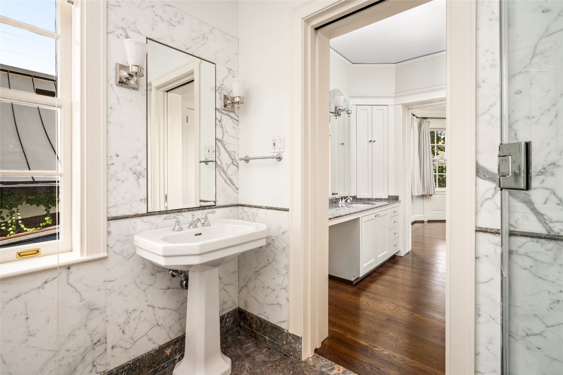 Marble lined bathroom with a pedestal sink and access to the adjoining bedroom and vanity area.