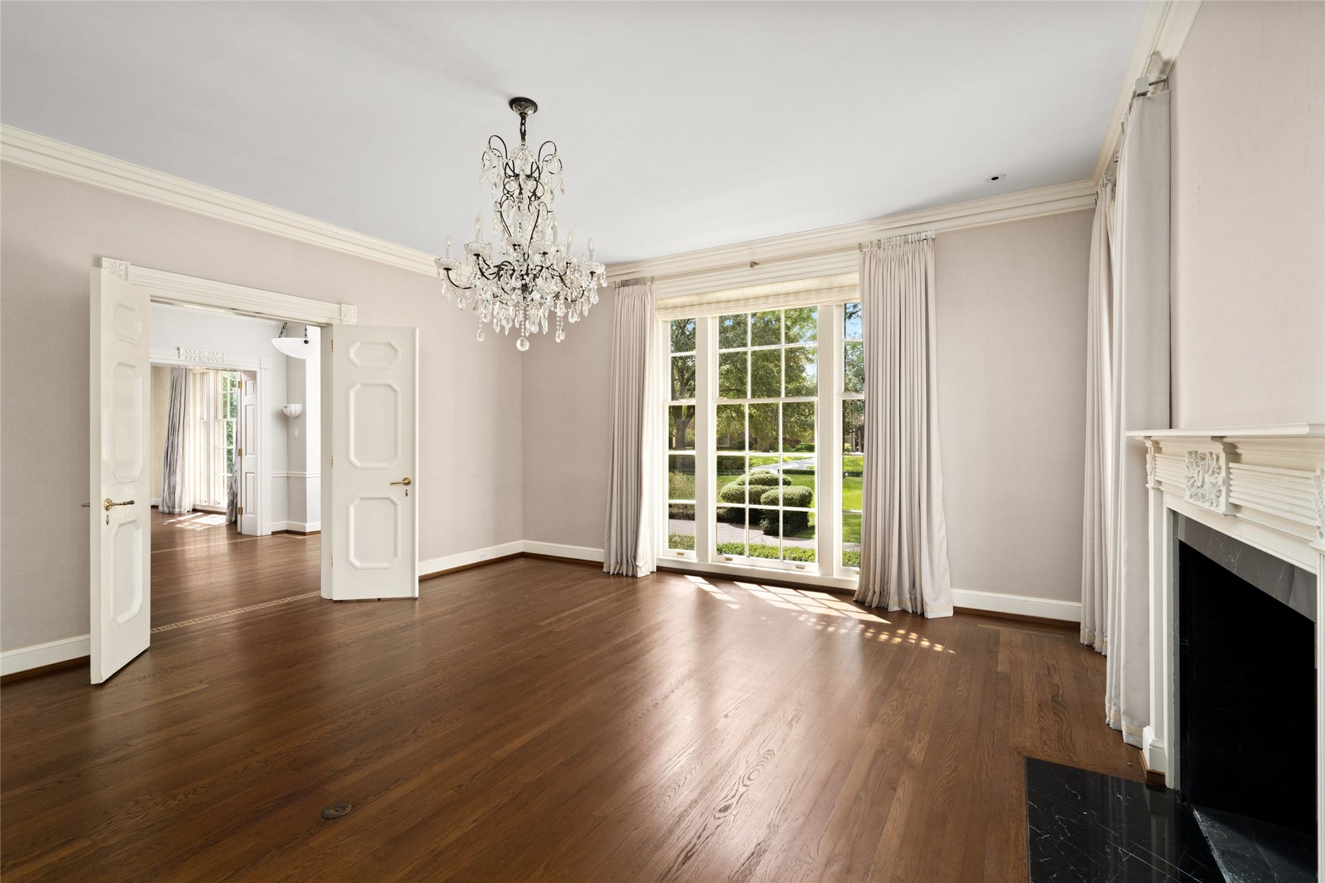 Formal Dinning room with a stunning chandelier, rich hardwood floors, and large windows offering natural light and garden views, complemented by classic French doors and a decorative fireplace.