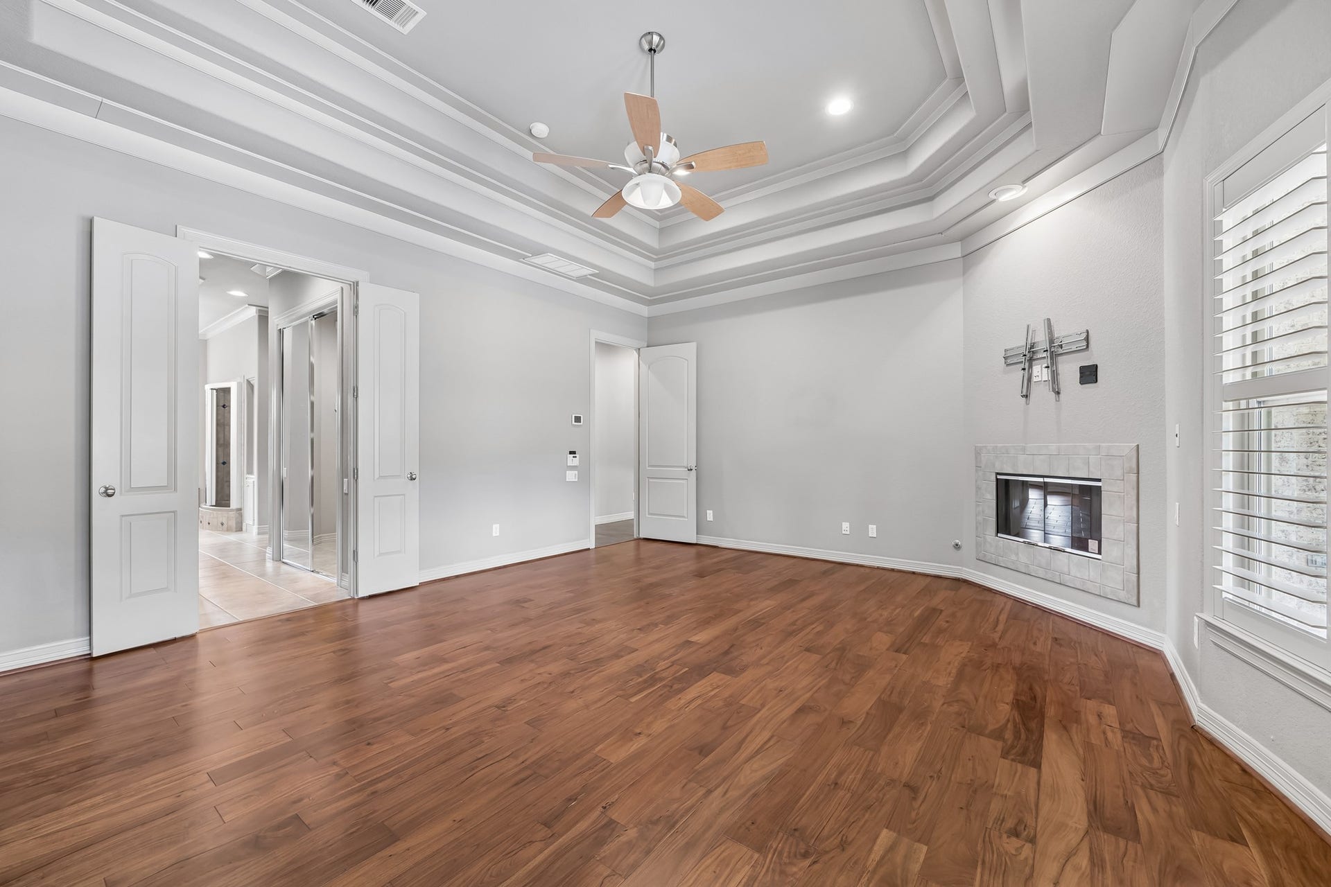 Spacious Primary Bedroom featuring elegant tray ceilings, warm wood flooring, and a cozy corner fireplace, accented by abundant natural light through plantation shutters.