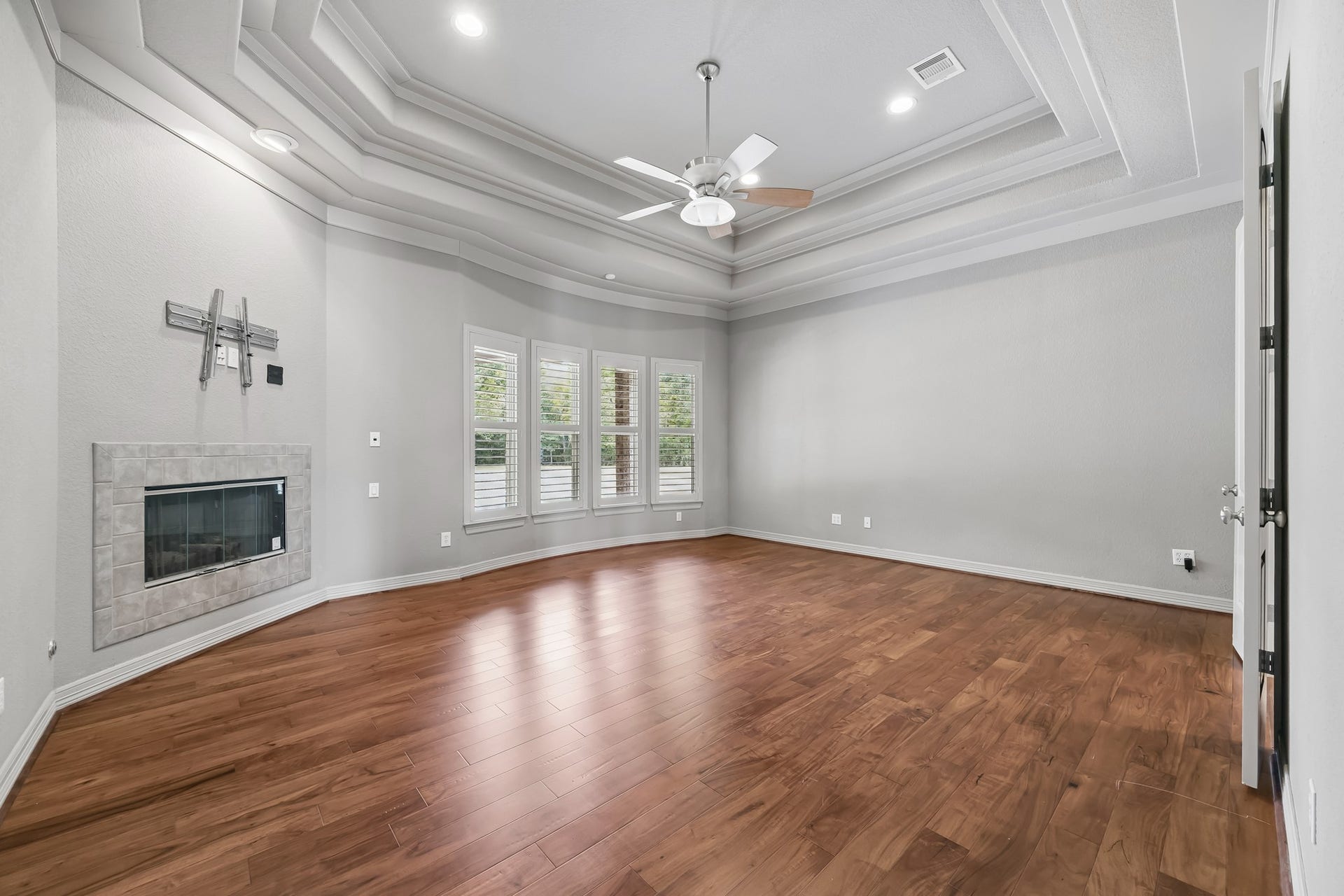 Spacious Primary Bedroom with elegant tray ceiling, rich hardwood floors, and a cozy modern fireplace, perfect for relaxation.