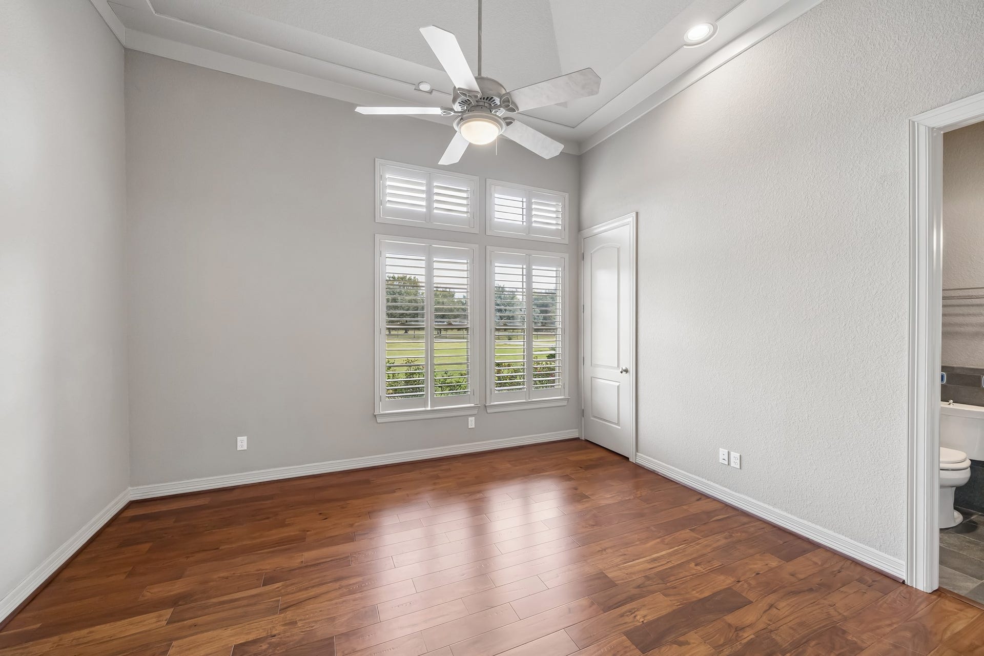 Bright ensuite bedroom with elegant wood flooring, large windows with plantation shutters, and a ceiling fan, offering a serene view of the outdoors.