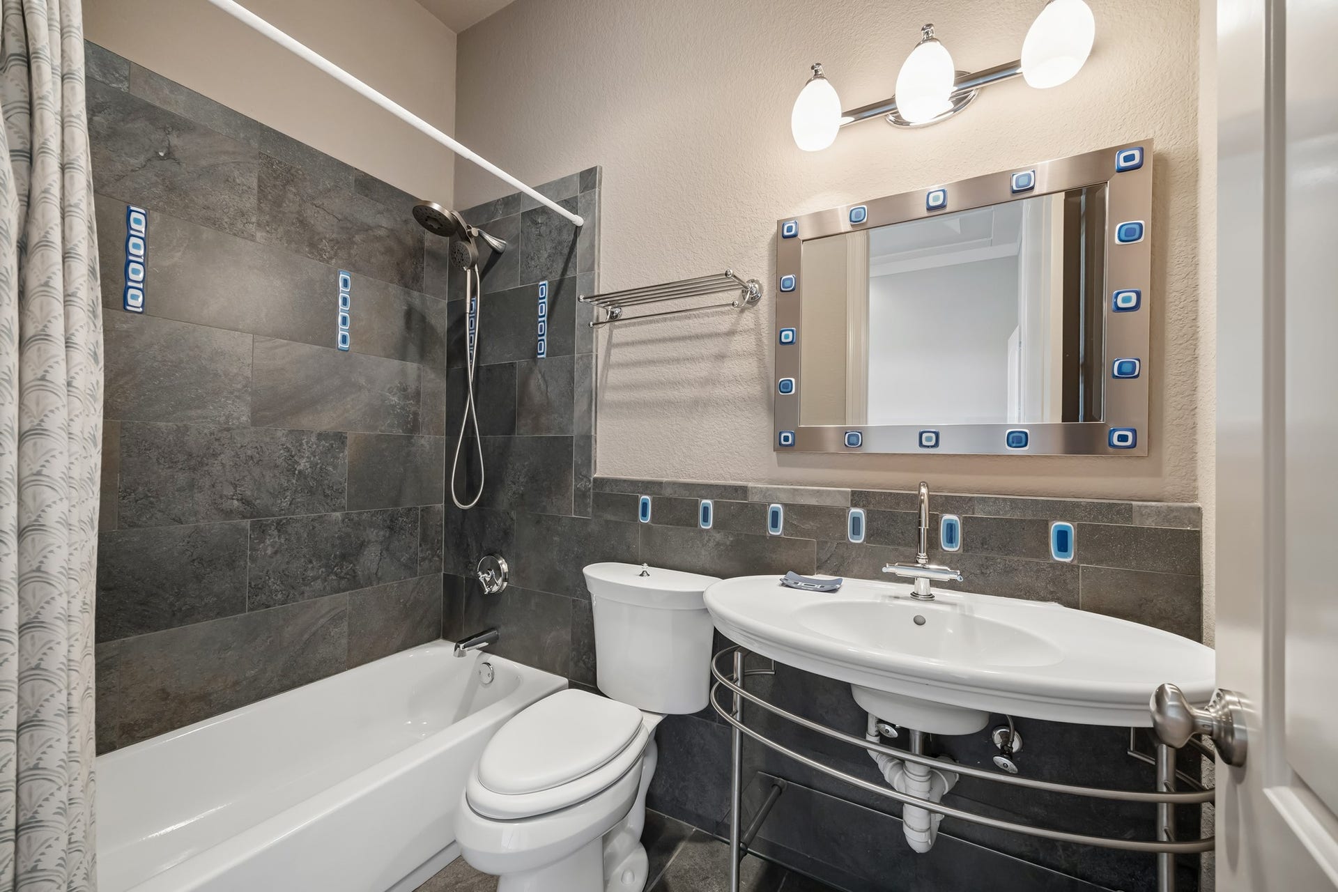 Modern bathroom with sleek dark tile, a stylish vanity, and a full bathtub, complemented by a unique mirror and contemporary lighting.