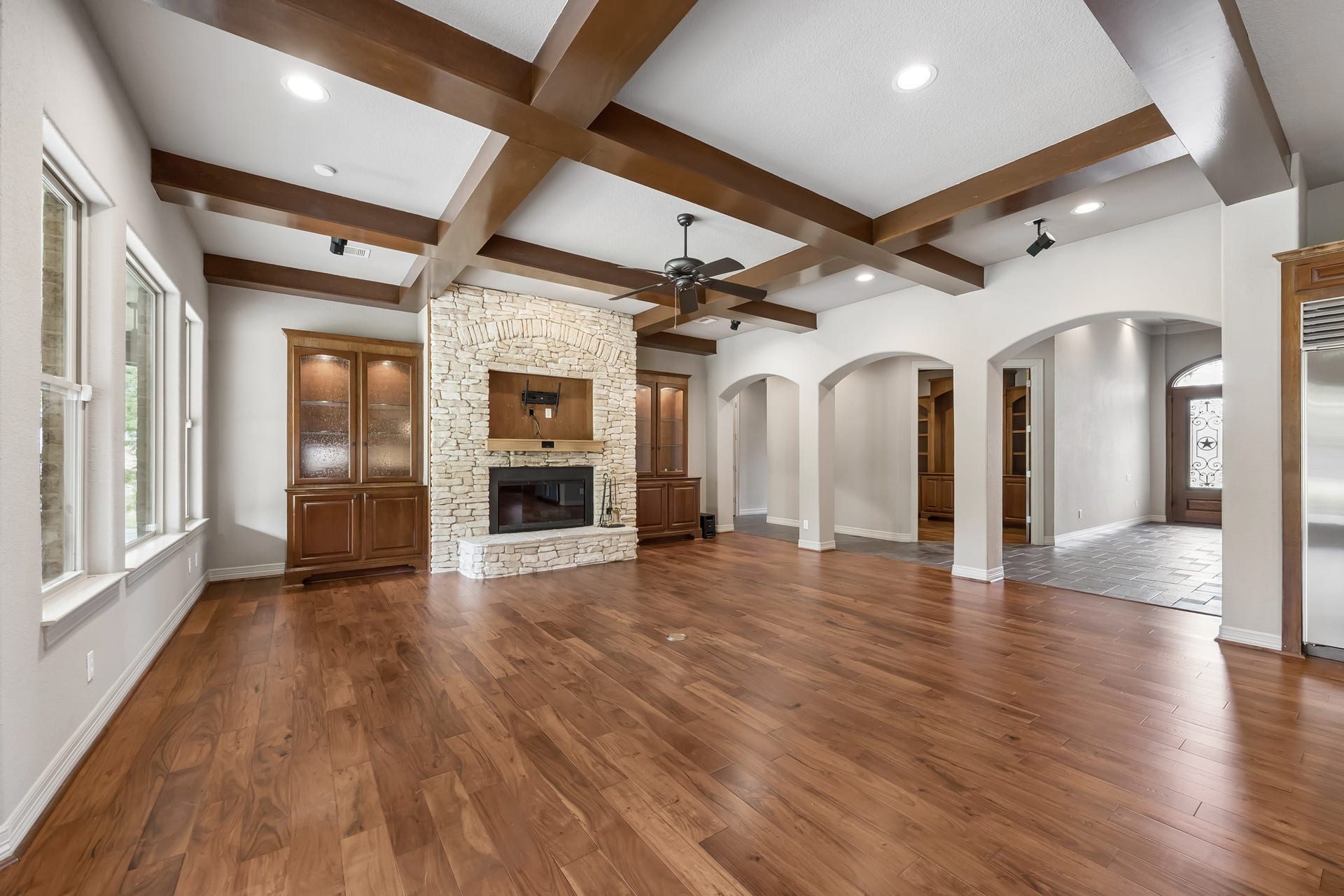 Spacious living area featuring rich hardwood floors, a striking stone fireplace, and elegant wood beam ceilings, complemented by built-in cabinetry and ample natural light.