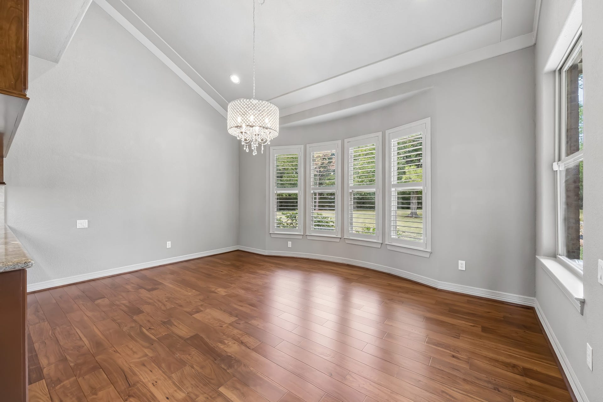 Elegant dining area featuring rich hardwood floors, a stunning chandelier, and large windows with plantation shutters overlooking the serene outdoors.