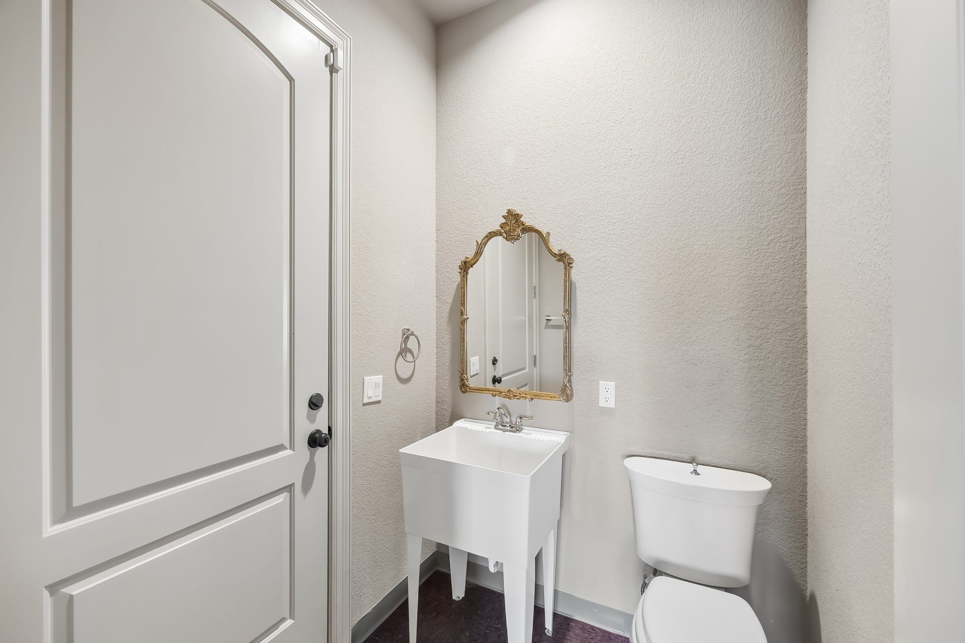 Half-bathroom featuring a classic pedestal sink with a gold-framed mirror and modern fixtures, set against soft, neutral walls.  Door exits to the garage.