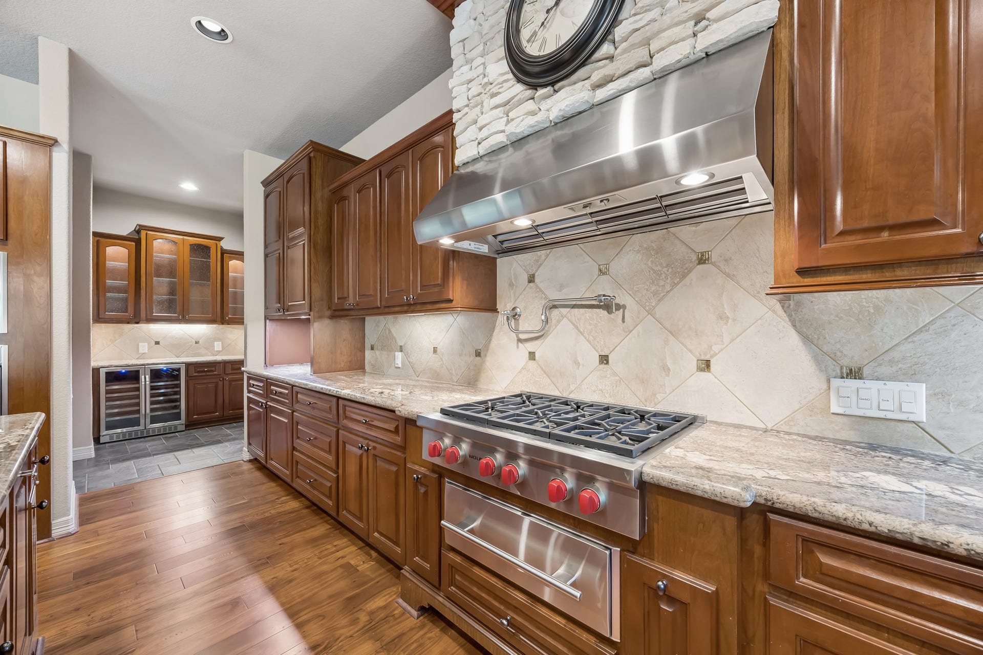 Elegant kitchen featuring rich wooden cabinetry, a gourmet gas range with a stylish hood, and a sleek backsplash. The adjacent butler's pantry offers additional storage and a wine cooler, perfect for entertaining.