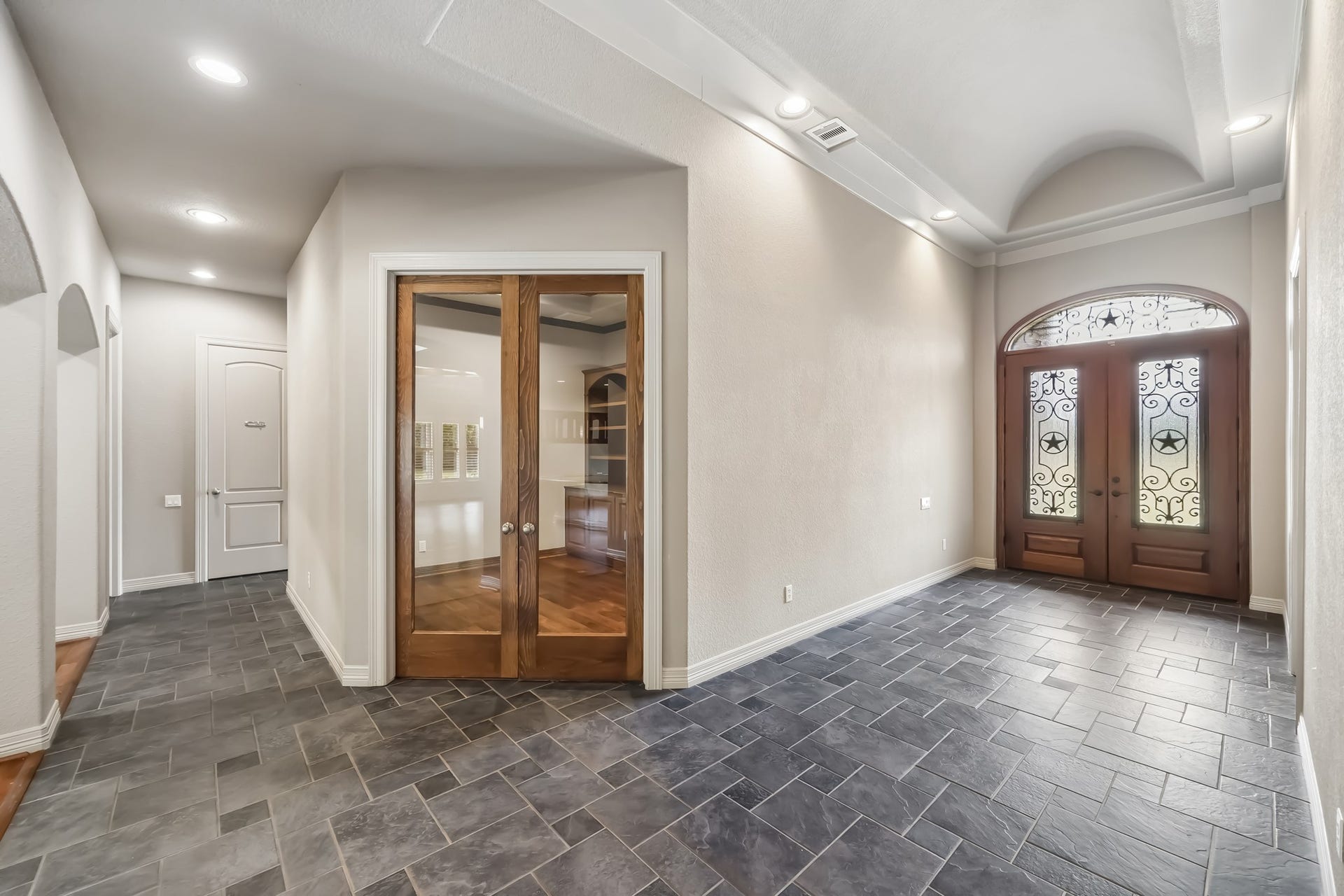 Elegant entryway featuring double wooden doors with decorative glass panels, set against sleek tile flooring and complemented by recessed lighting and high ceilings. The office is located behind the double doors with built in custom shelves.
