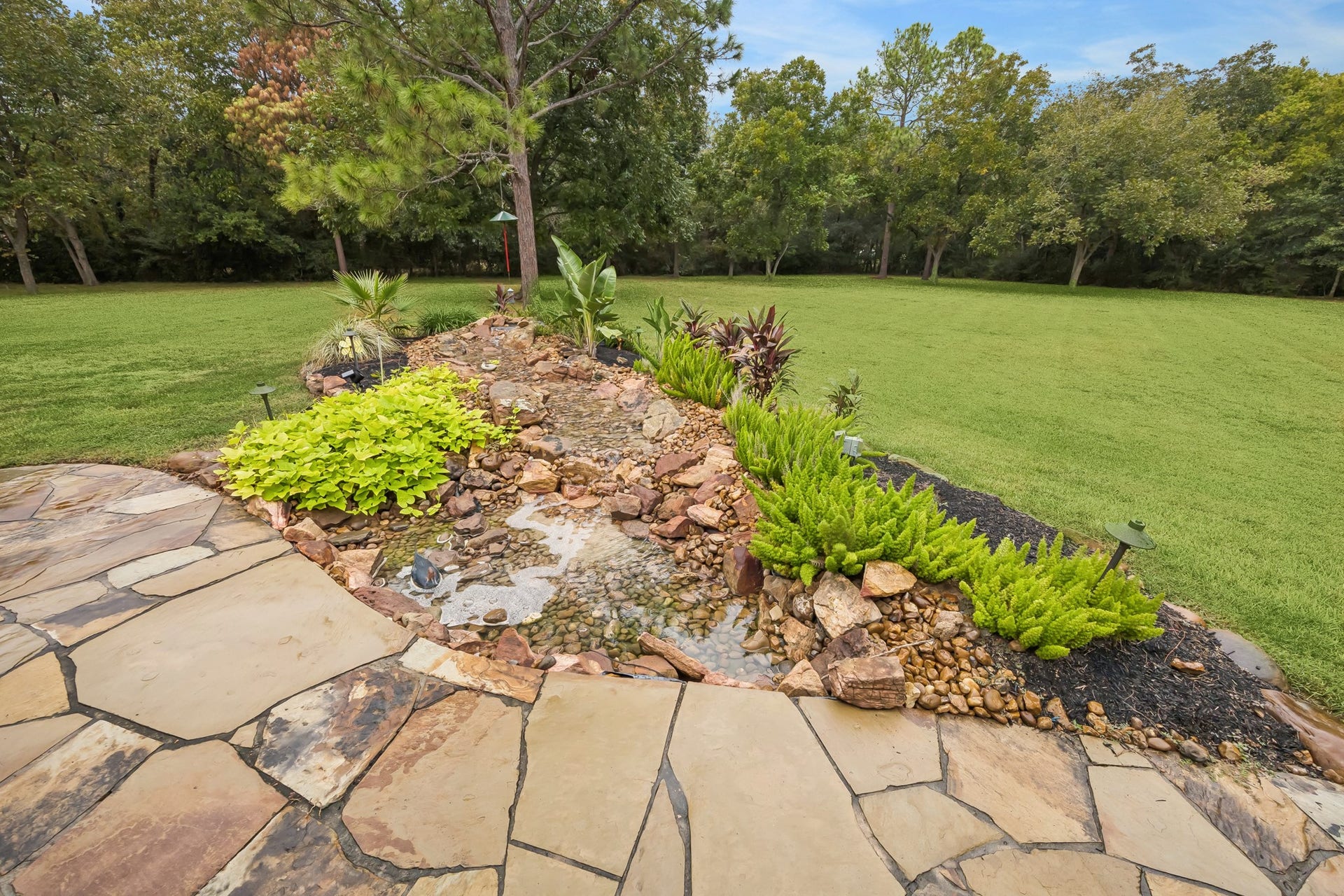 Discover a serene backyard oasis featuring a beautifully landscaped garden with a tranquil rock-lined water feature, bordered by lush greenery and a spacious lawn.