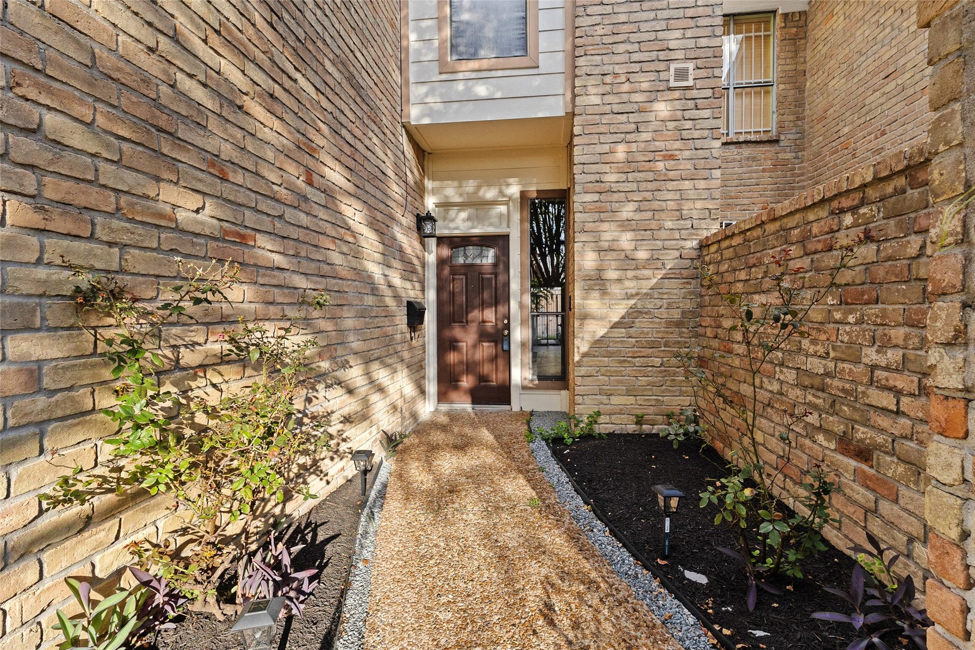Your welcoming front entrance features classic brick walls, landscaped garden beds, decorative pathway lighting, and a rich wood-tone front door with a glass sidelight, creating a warm and inviting first impression.