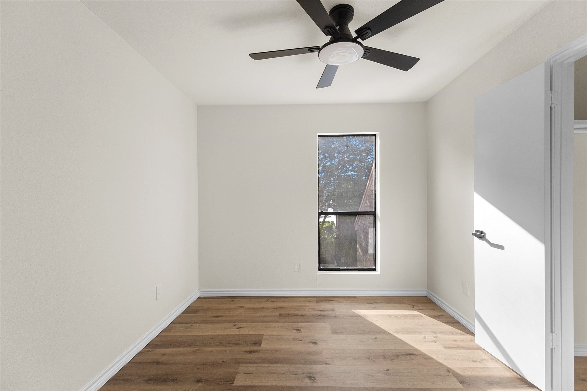 Your third bedroom offers warm wood-look flooring, neutral walls, a large window providing natural light, and a modern ceiling fan with integrated lighting.
