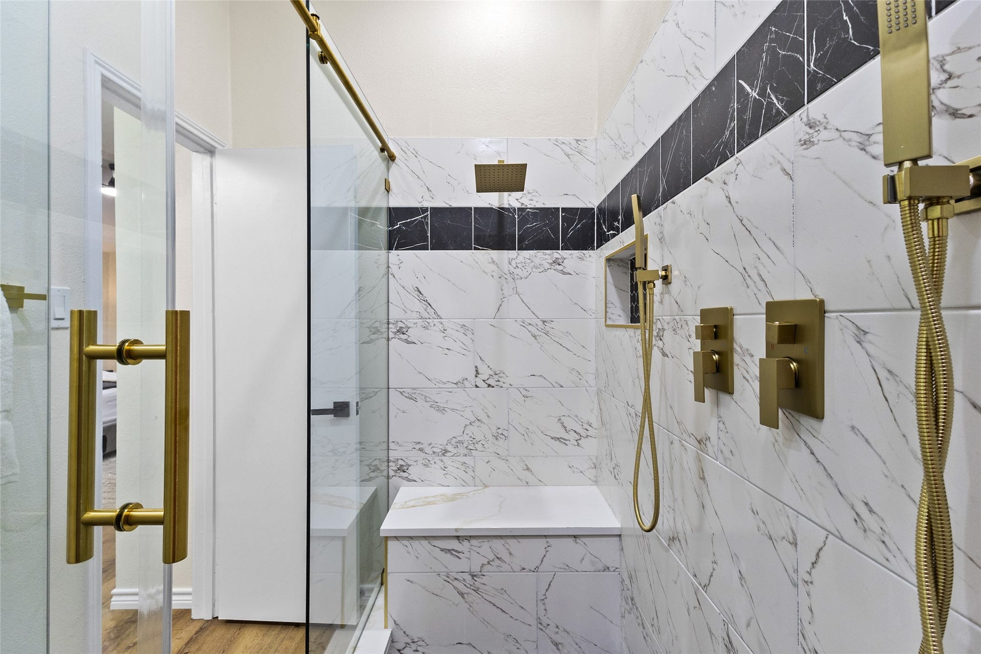 Your massive primary bathroom shower features elegant marble-look tile, bold black accent detailing, brushed gold fixtures, a rainfall showerhead, handheld shower, and a cbuilt-in bench.