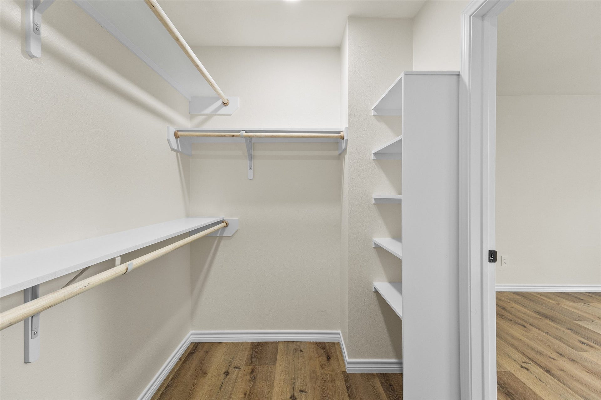 Your spacious walk-in closet for the secondary bedroom showcases a well-planned design with multiple hanging rods, convenient built-in shelving, and ample room to neatly arrange clothing, footwear, and personal belongings. Fresh white walls and matching storage features complement the warm wood-look flooring, creating a bright, polished, and functional space that combines style with practical everyday storage solutions.