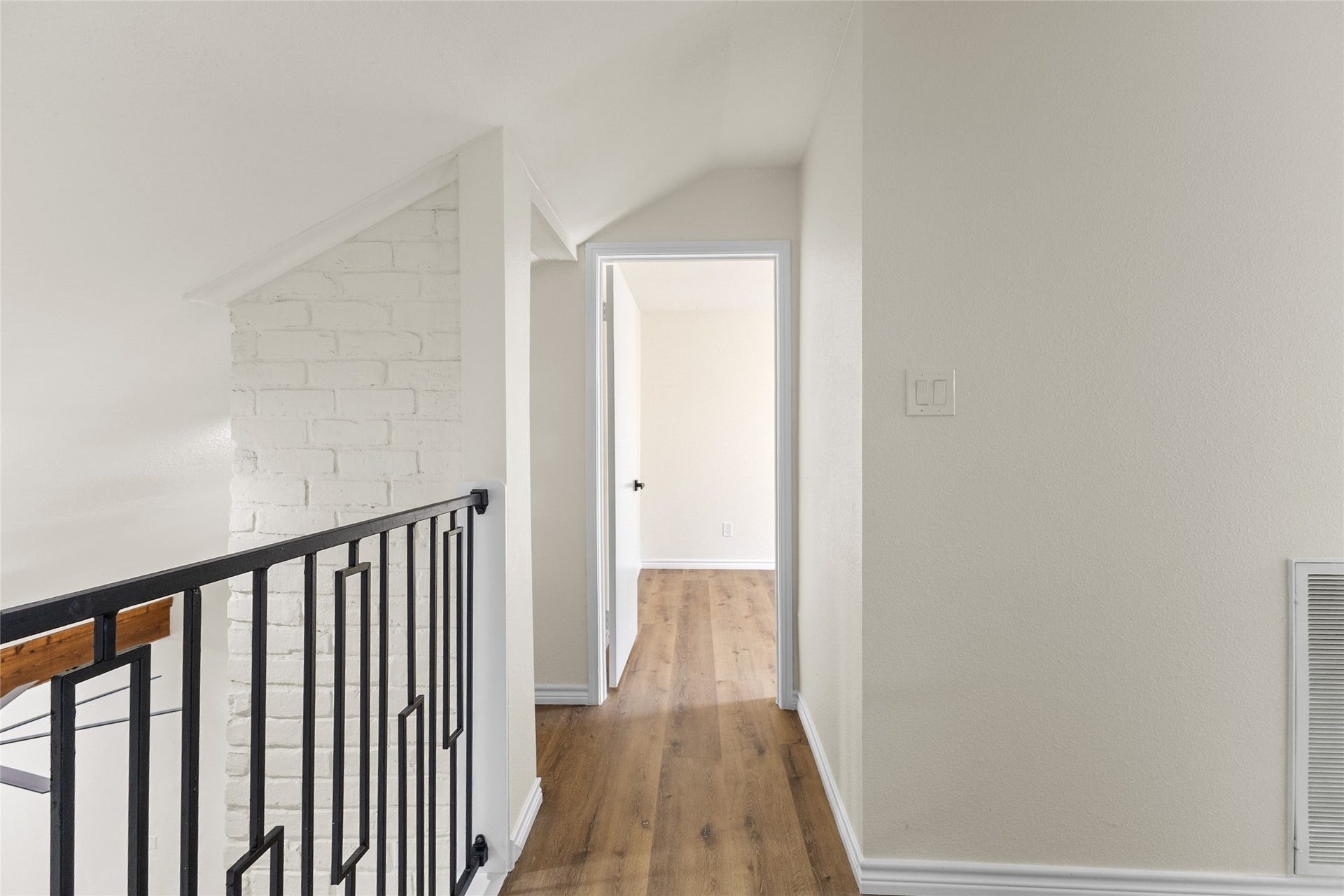 Your upstairs hallway showcases warm wood-look flooring, a sleek black metal railing, and a distinctive white-painted brick accent wall that adds texture and architectural character. The combination of modern finishes and contrasting details creates a stylish transition between the upper-level living spaces.