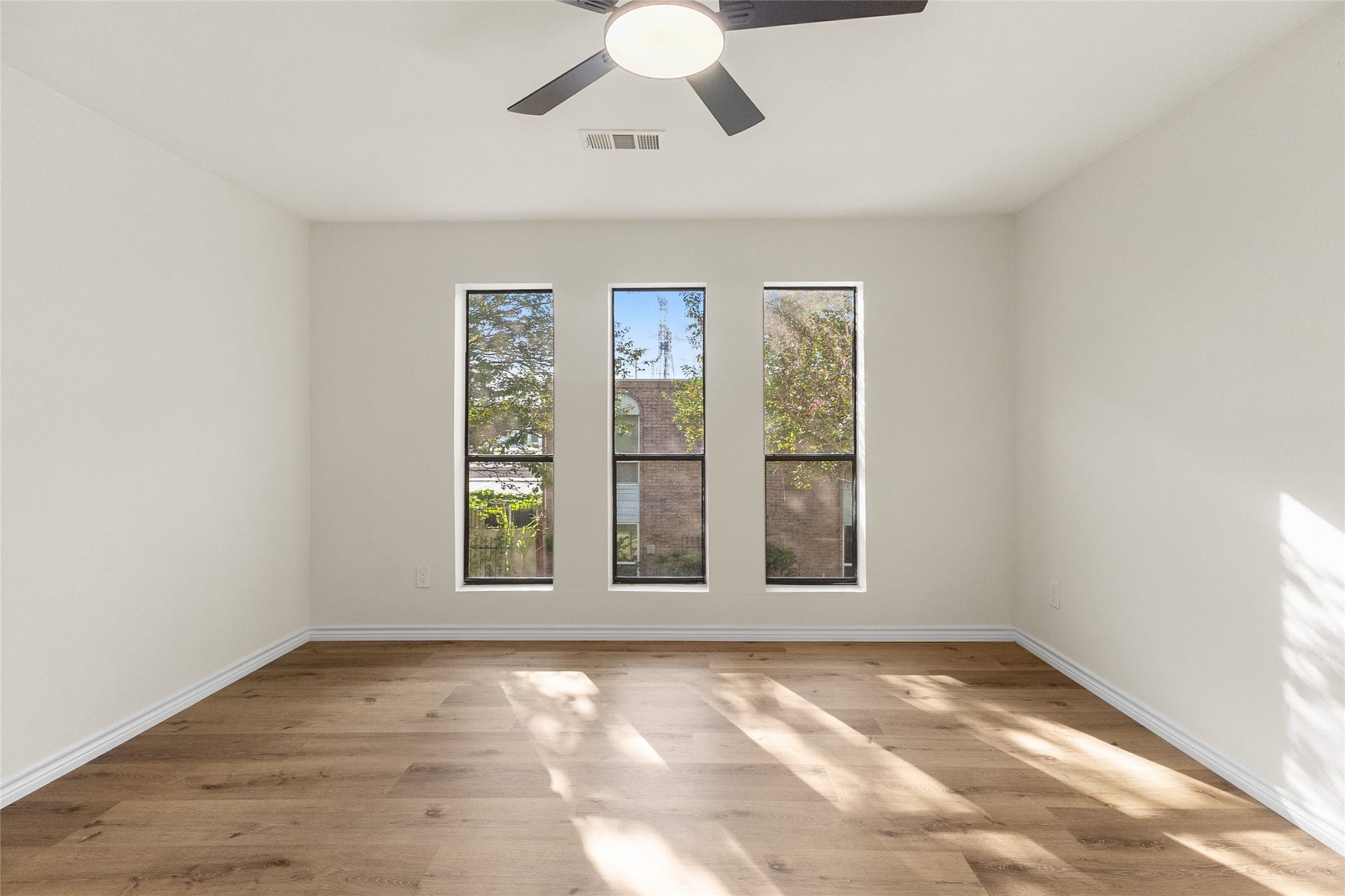 Upstiars, your bright and spacious secondary bedroom features three tall windows that fill the room with beautiful natural light, wood-look flooring, neutral walls, and a contemporary ceiling fan.